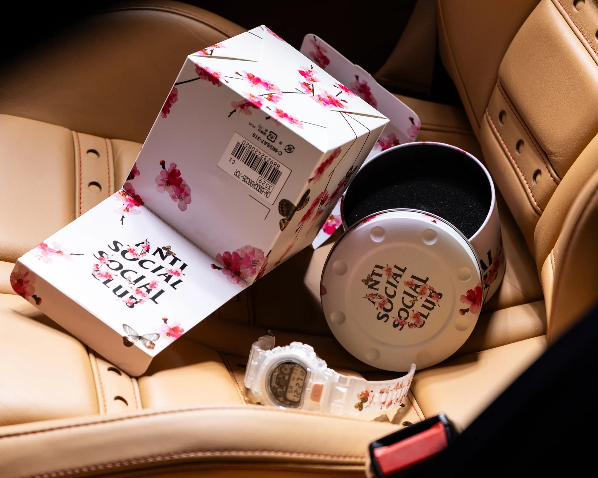 White floral-patterned Anti Social Social Club watch with matching round box and packaging on tan leather car seat.