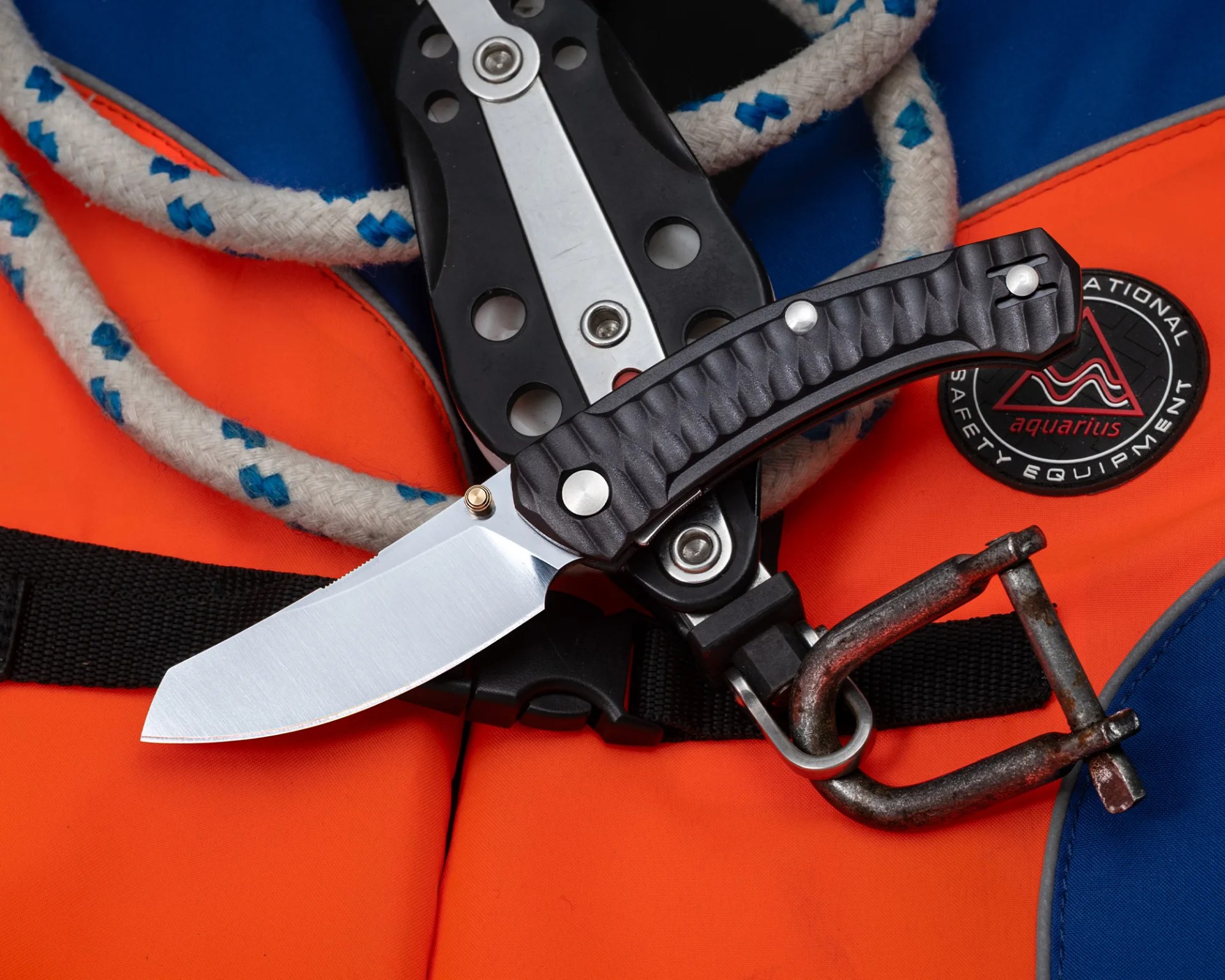 Folding knife with textured black handle and silver blade resting on orange and blue safety equipment with rope and metal shackle.