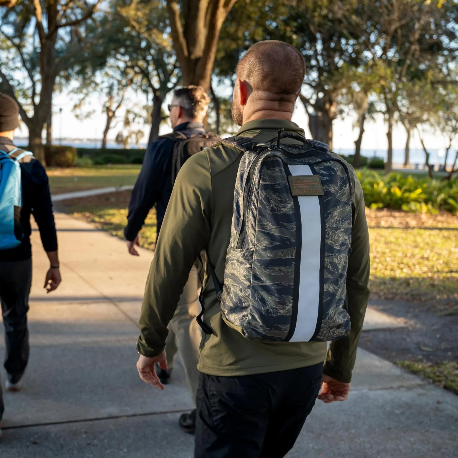 Camouflage-patterned backpack with a vertical white stripe worn by a person walking on a tree-lined sidewalk.