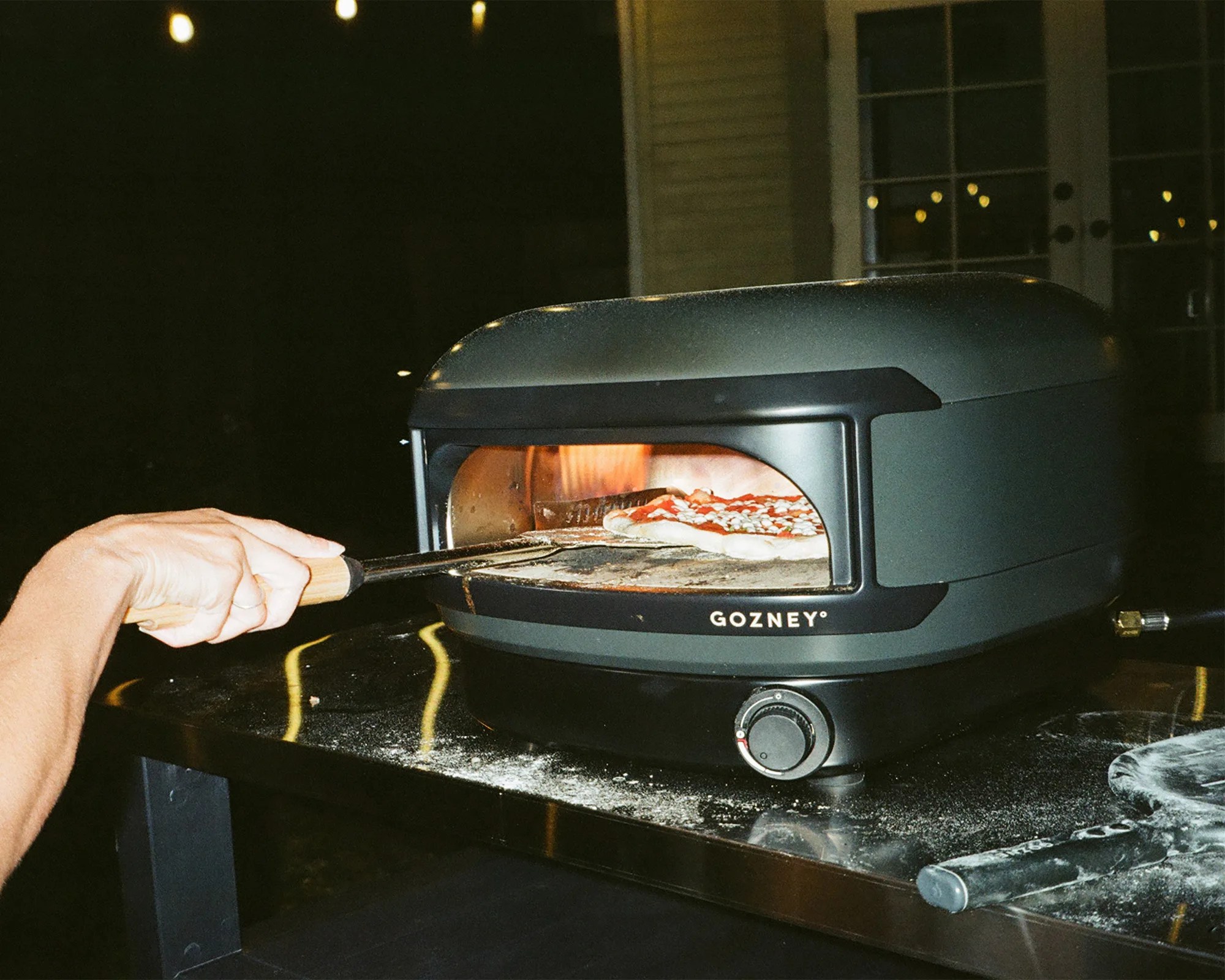 Black Gozney pizza oven on a metal table with a pizza peel holding a pizza inside the oven.