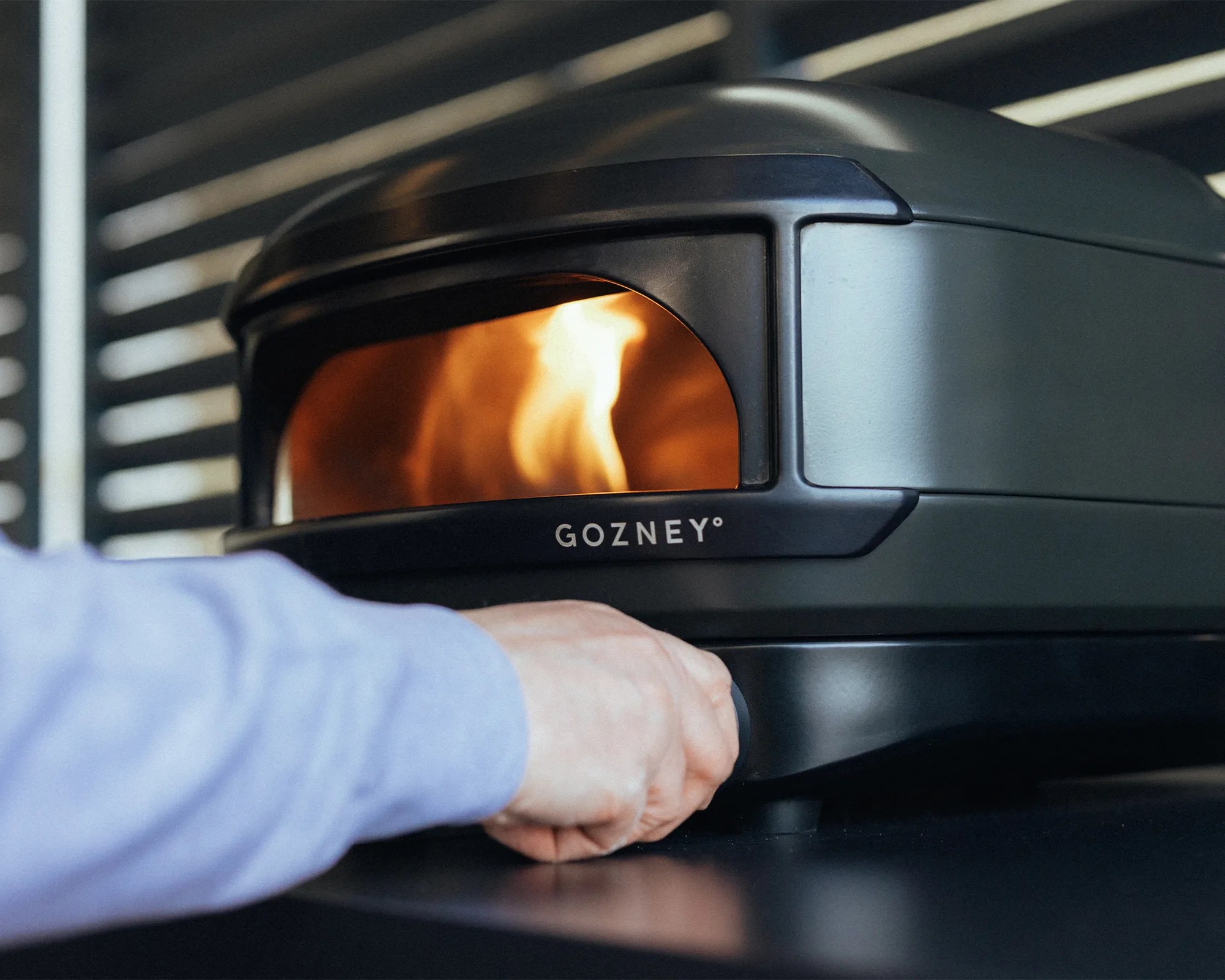 Hand adjusting a knob on a black and gray Gozney outdoor pizza oven with visible flames inside.