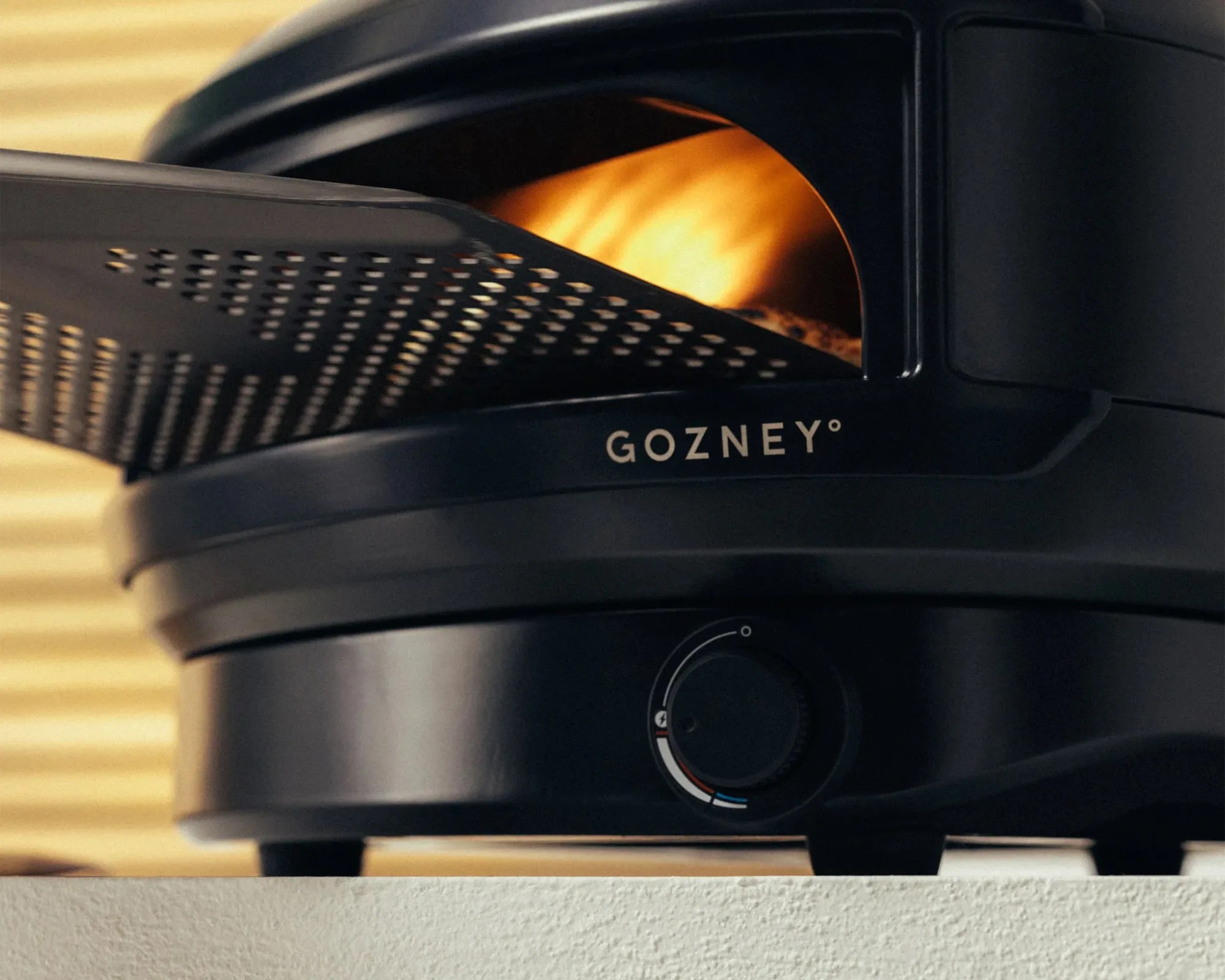 Black Gozney countertop pizza oven with open perforated door showing flames inside.