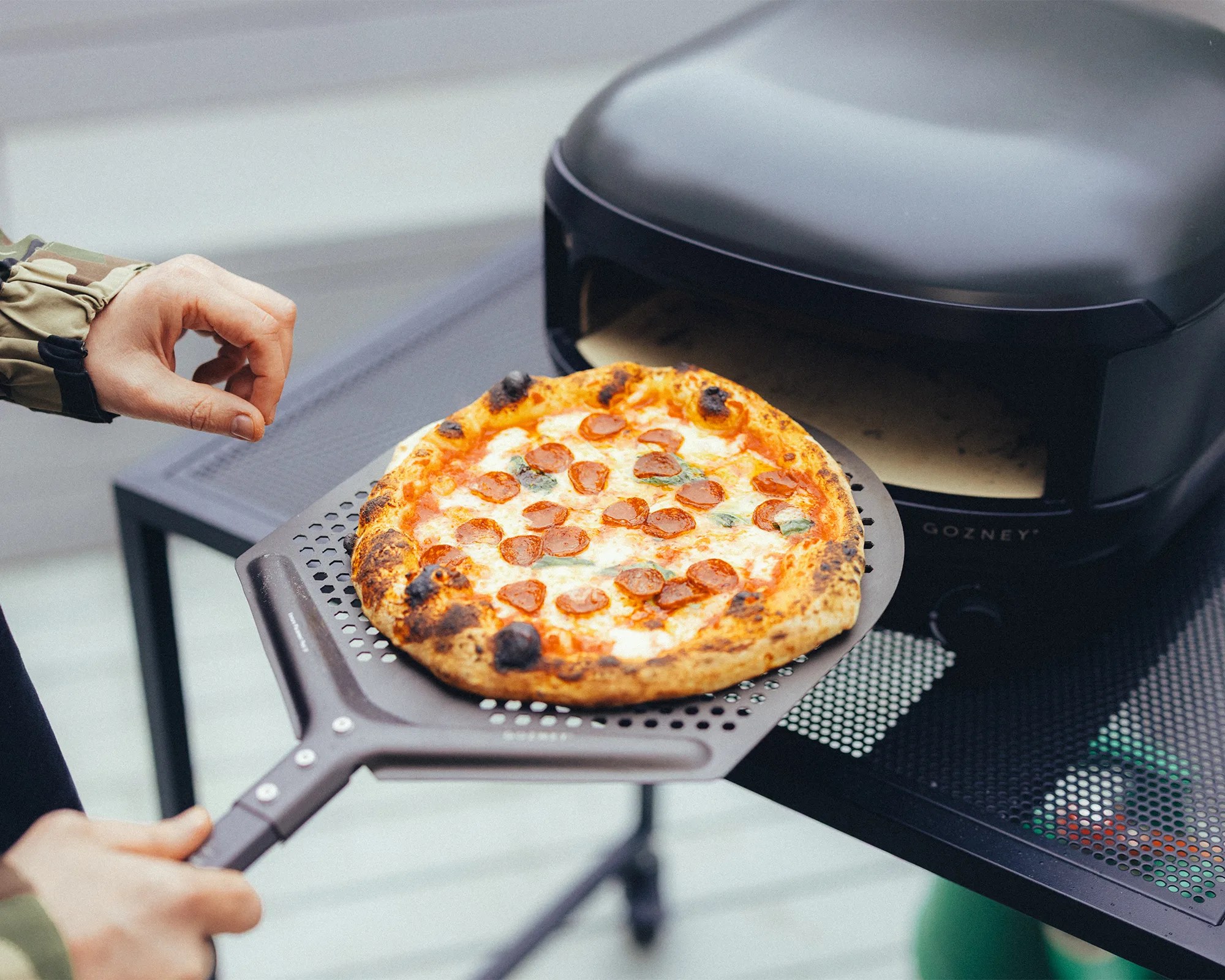 Pepperoni pizza with charred crust on a black perforated pizza peel near a black Gozney pizza oven.
