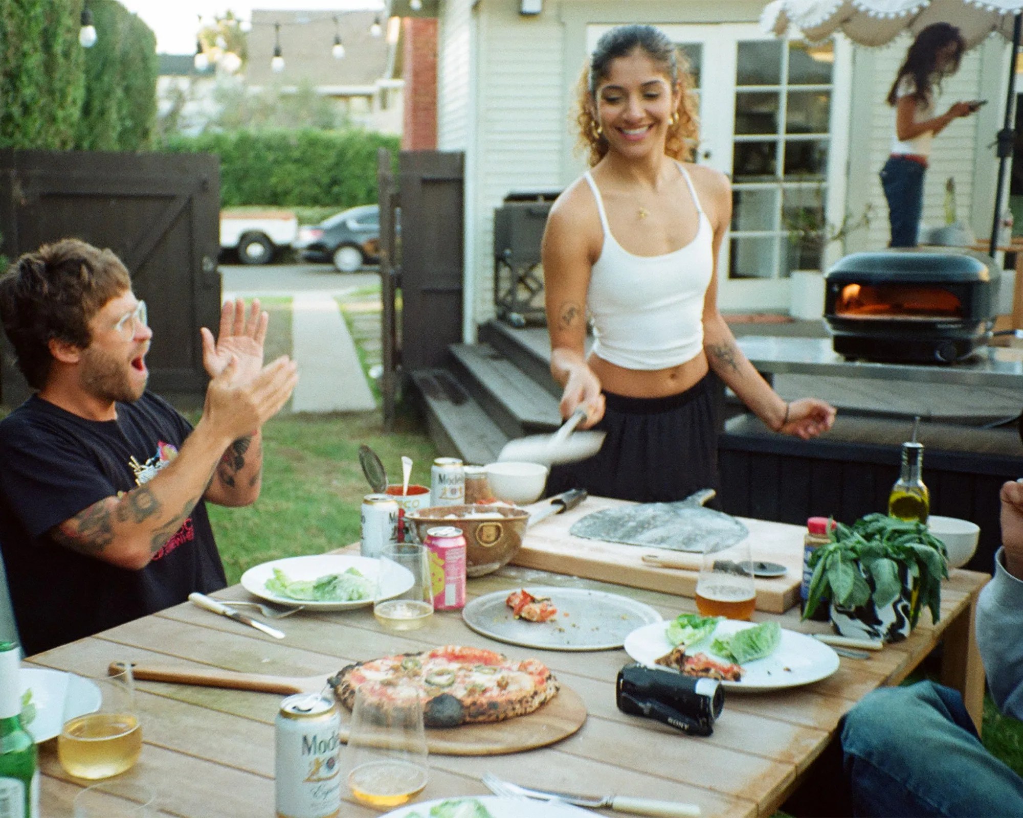 Outdoor wooden table with pizza, drinks, and a woman serving food while a man claps nearby.