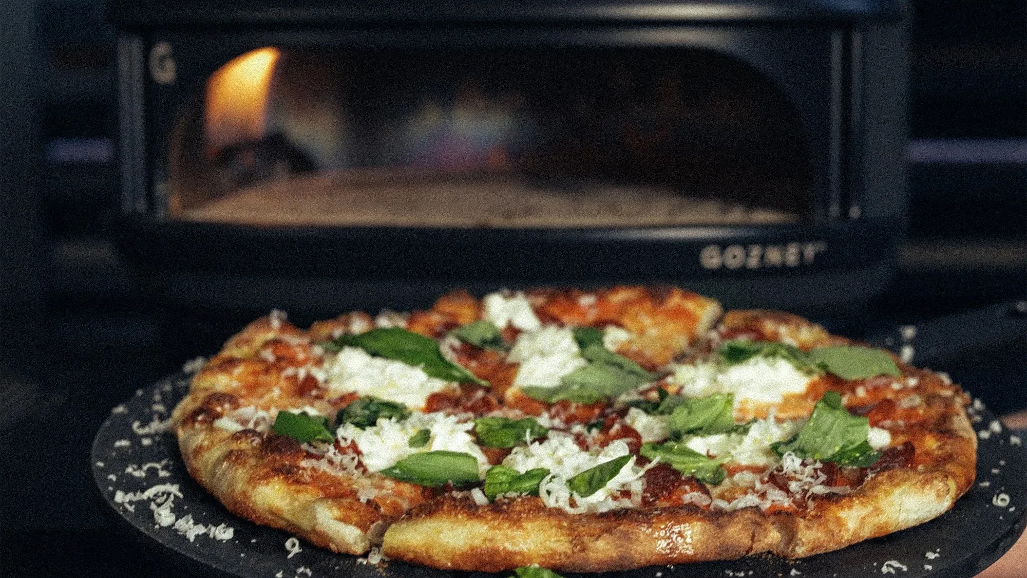Thin-crust pizza topped with ricotta cheese and fresh basil leaves on a black pizza peel in front of a pizza oven.