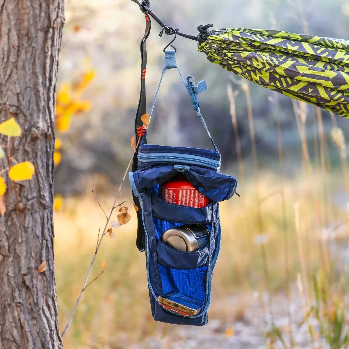 Blue hanging gear organizer with a red canister, metal cup, and phone inside, attached to a green patterned hammock.