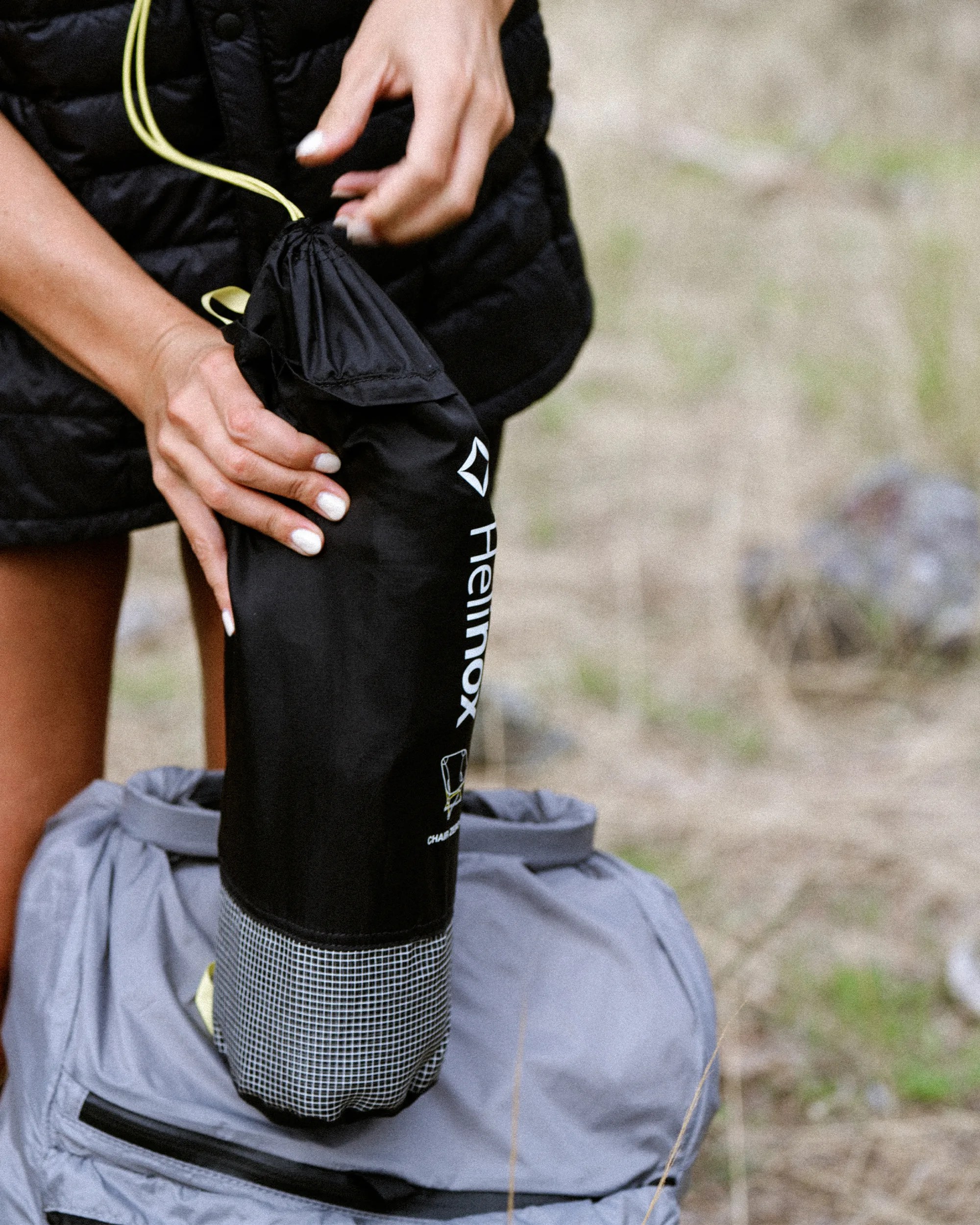 Person holding a black Helinox drawstring bag above a gray backpack outdoors.