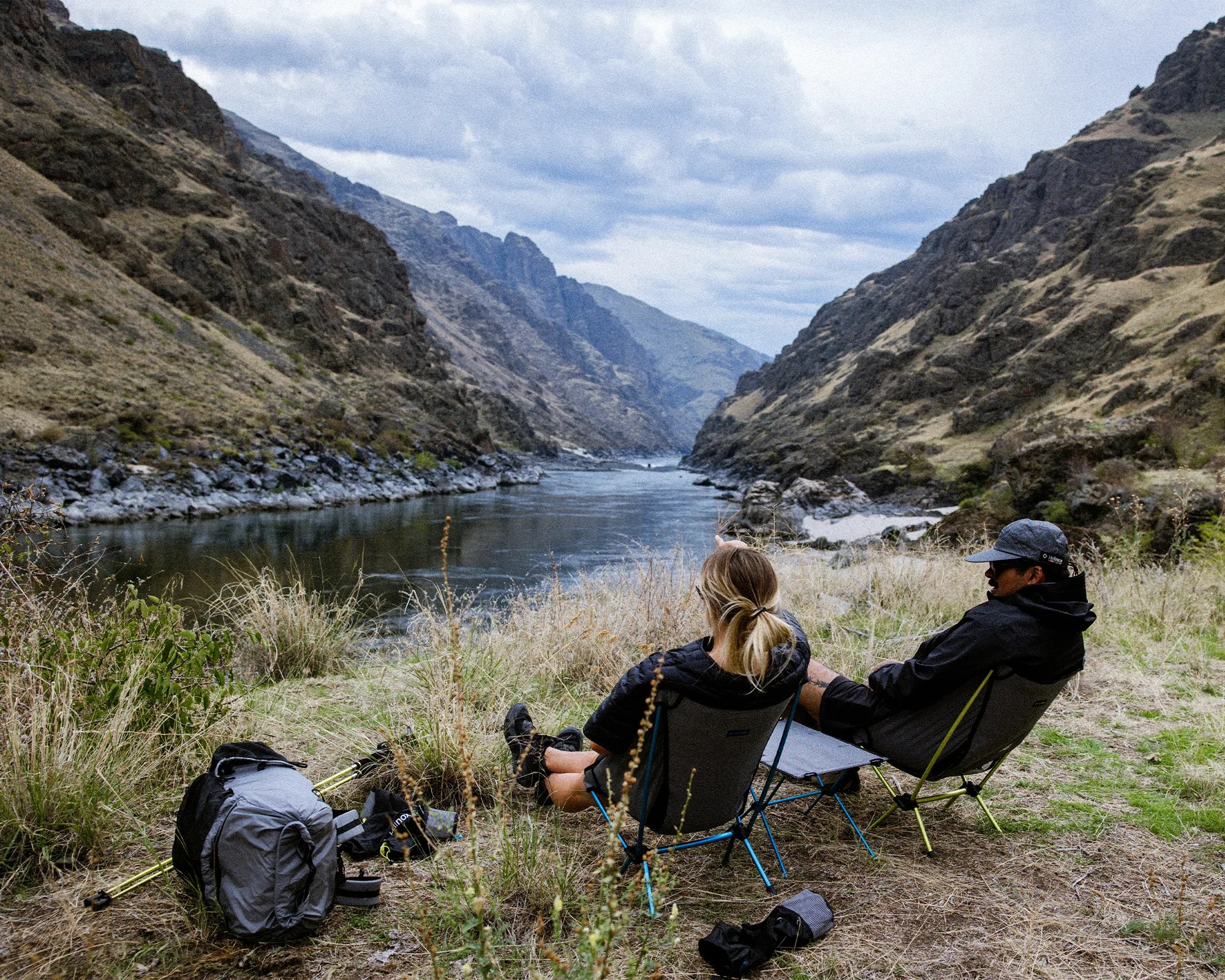 Two people sitting in portable camping chairs by a river in a mountainous landscape with backpacks and hiking poles nearby.
