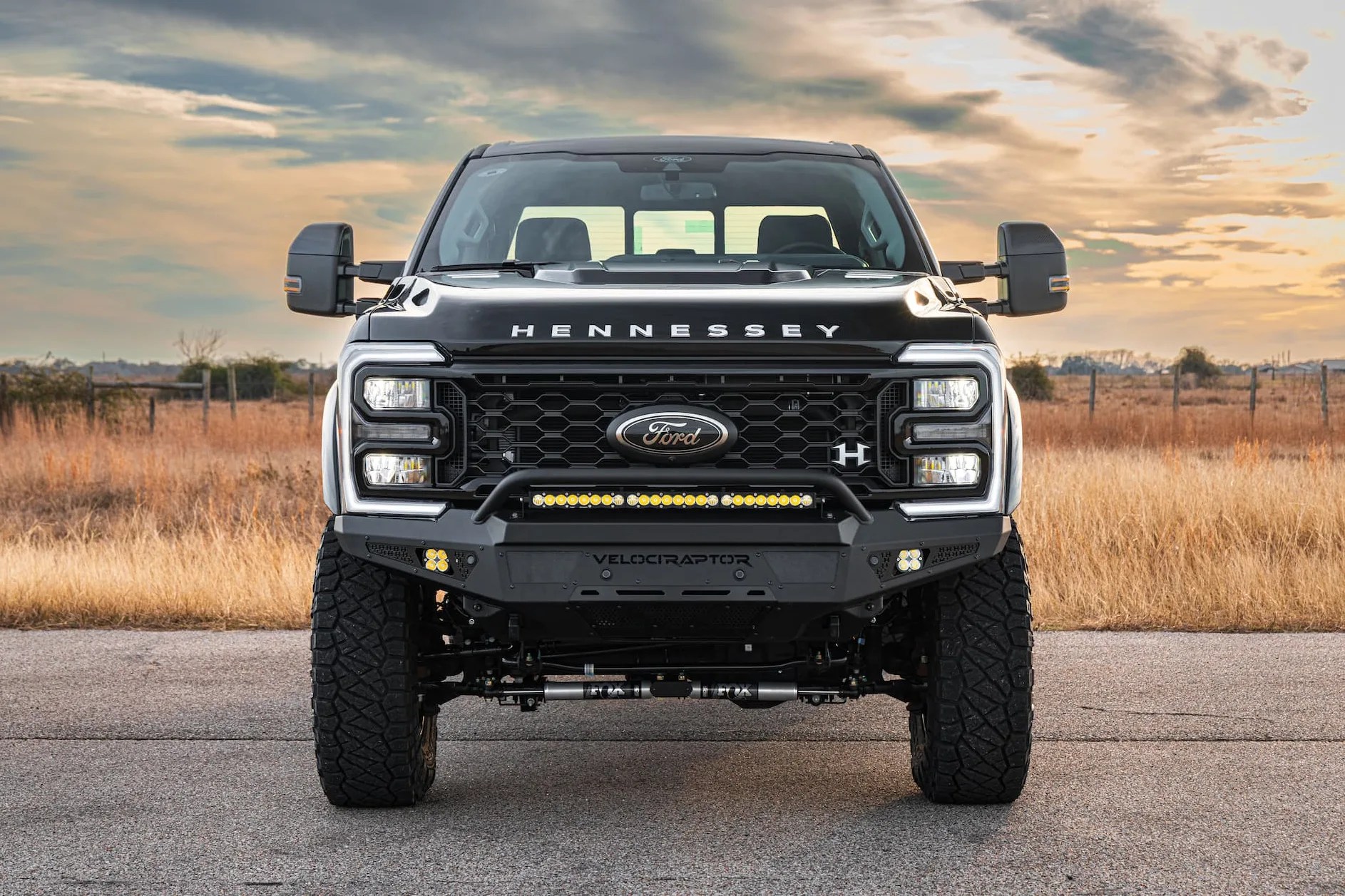 Front view of a black Hennessey VelociRaptor Ford truck with large off-road tires and LED light bar on a rural road.