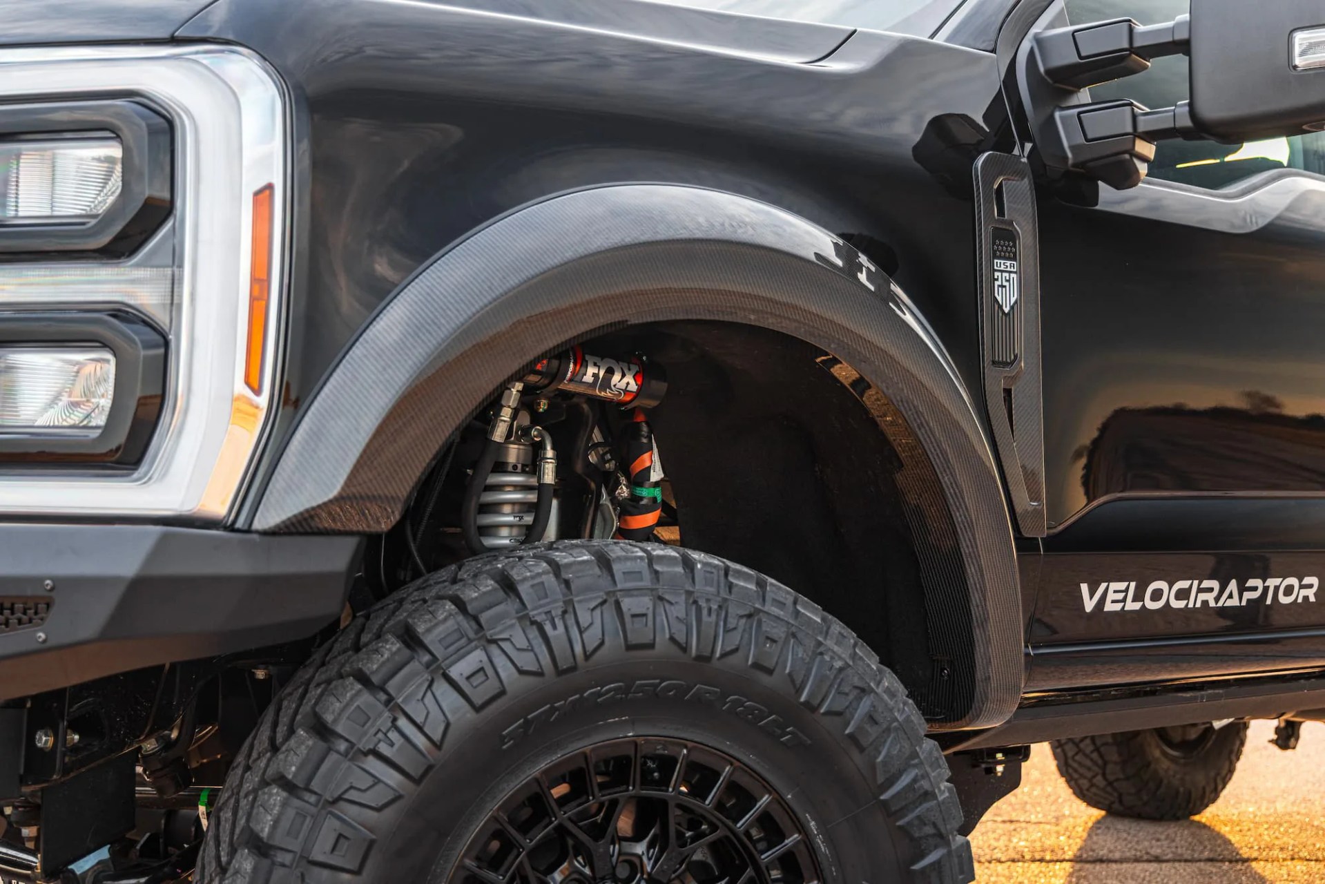 Close-up of a black truck's front wheel with large off-road tire, carbon fiber fender flare, and "Velociraptor" decal on the door.