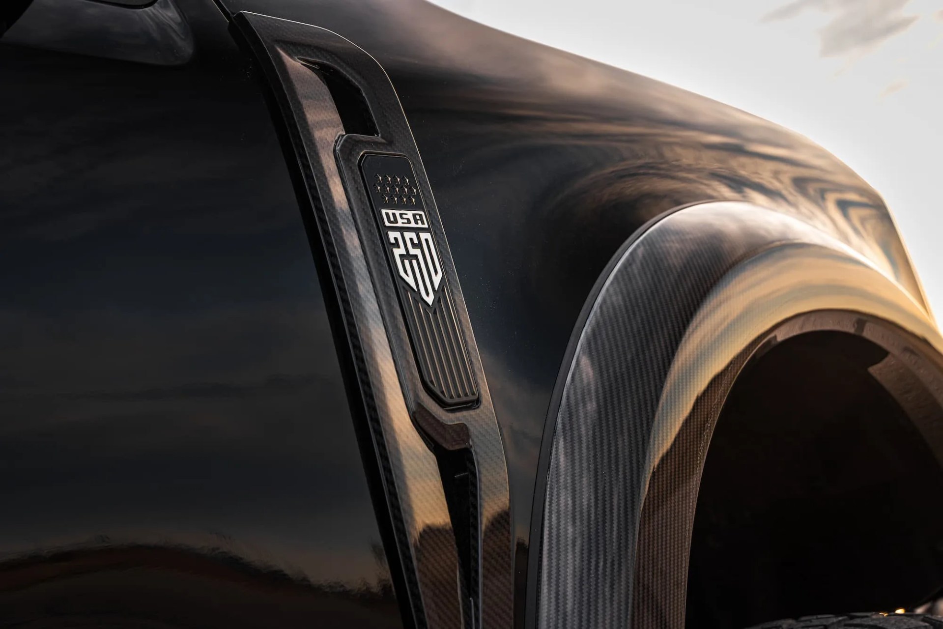 Close-up of a black vehicle fender with a carbon fiber trim and a "USA 250" emblem.
