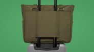Olive green fabric tote bag secured on the handle of a silver rolling suitcase against a green background.