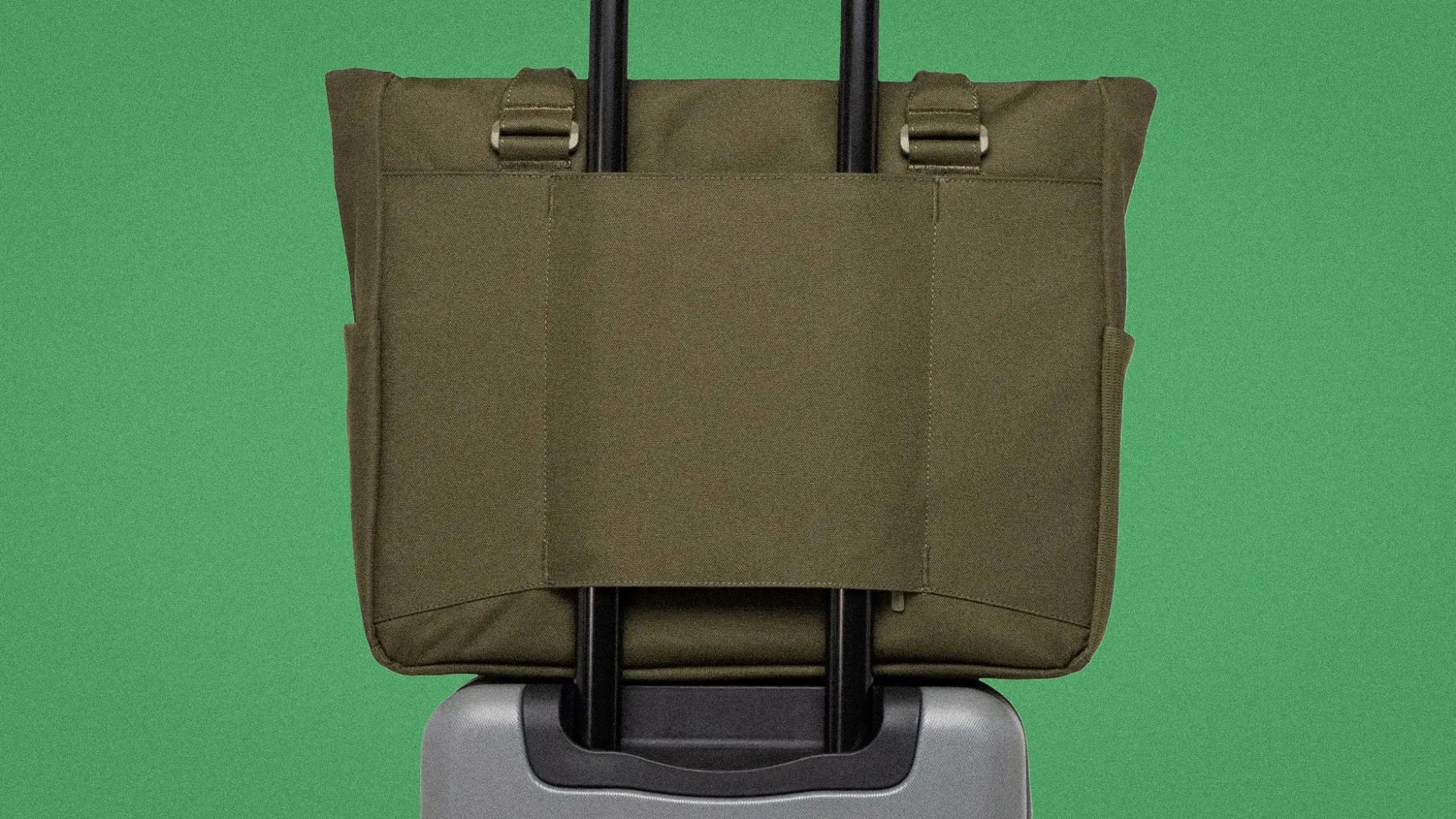 Olive green fabric tote bag secured on the handle of a silver rolling suitcase against a green background.
