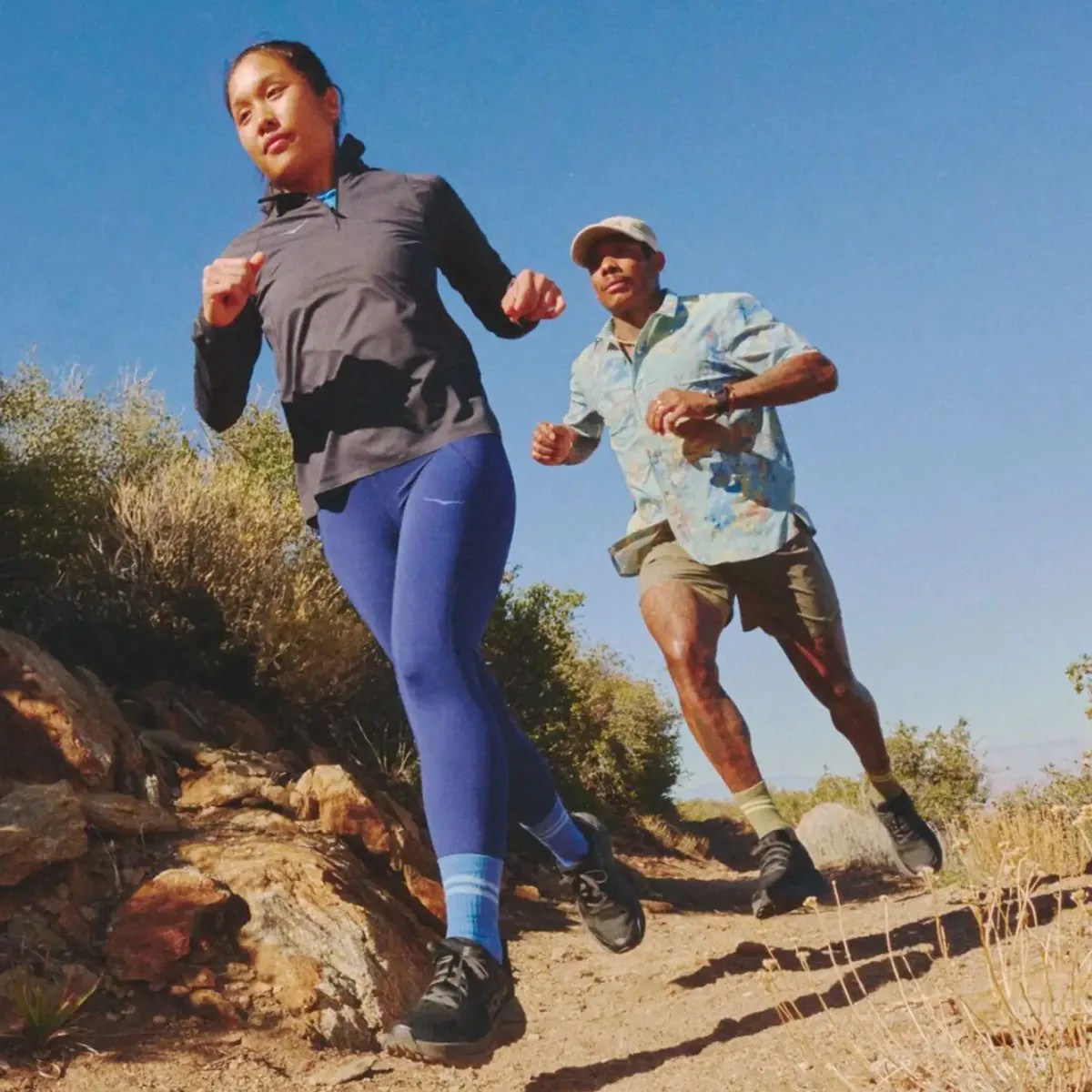 Two people trail running on a rocky dirt path under a clear blue sky.