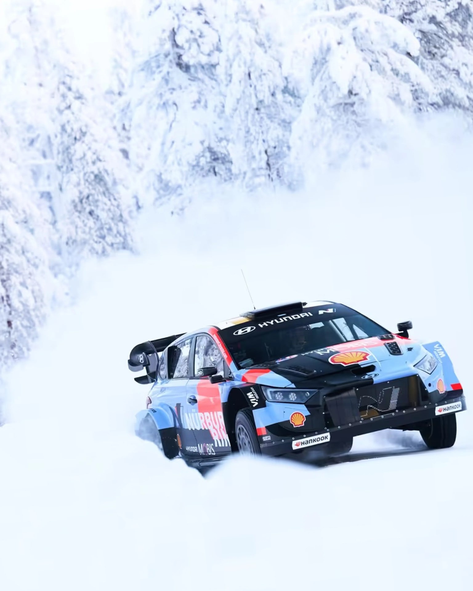 Blue and black Hyundai rally car with Shell and Hankook logos drifting on snowy road with snow-covered trees in background.