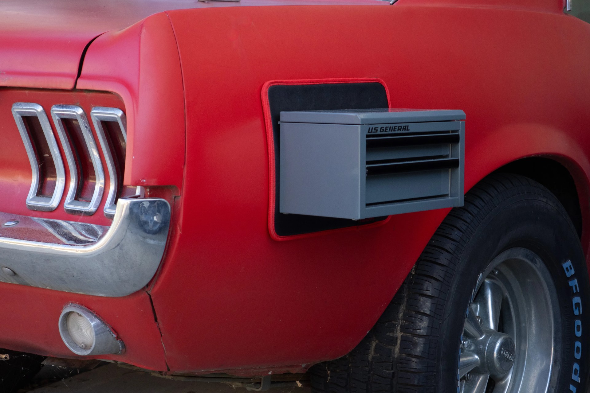 Gray US General tool chest mounted on the red rear quarter panel of a vintage car near the tire.
