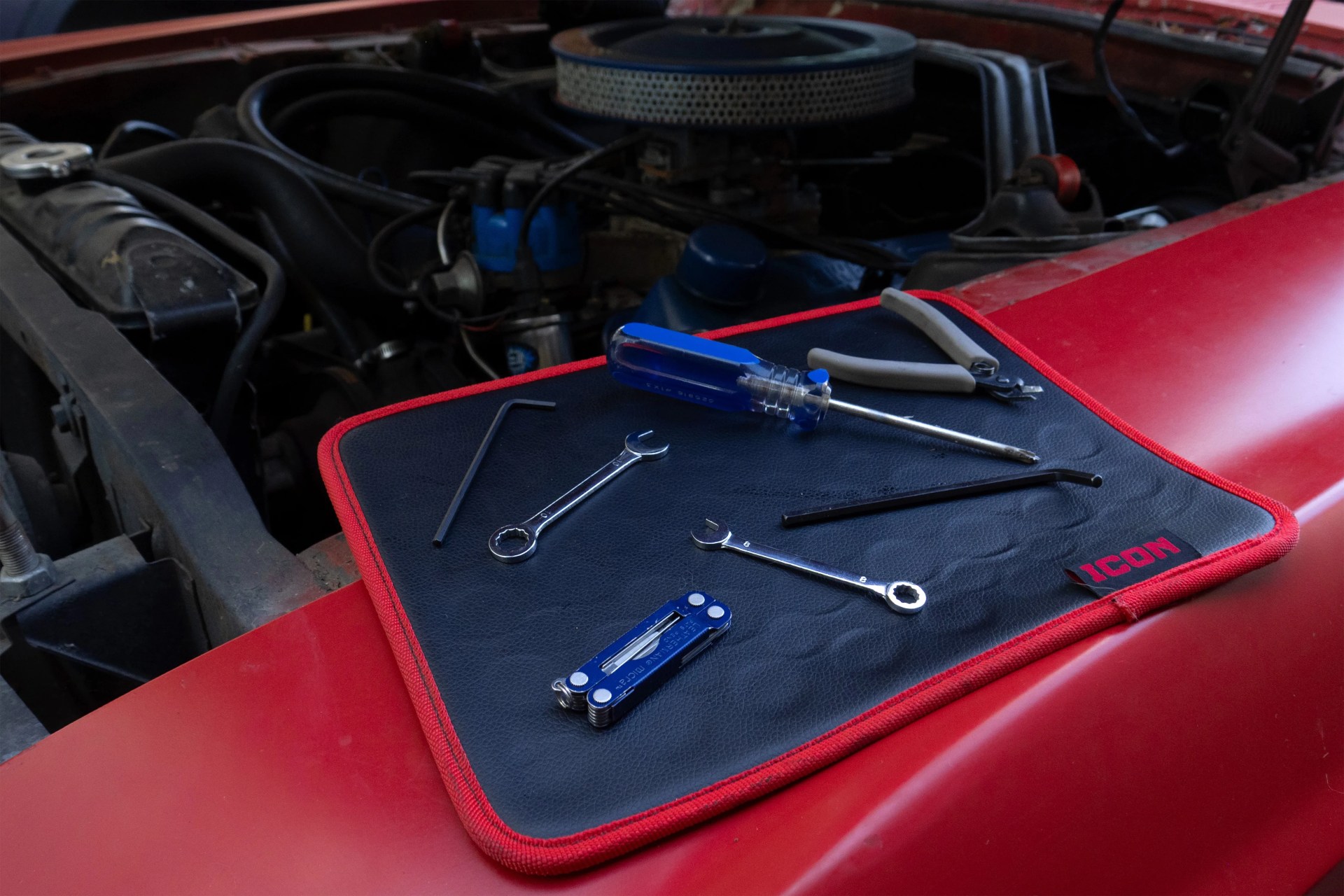 Black and red ICON tool mat on a red car hood with various hand tools including wrenches, screwdrivers, pliers, and hex keys.
