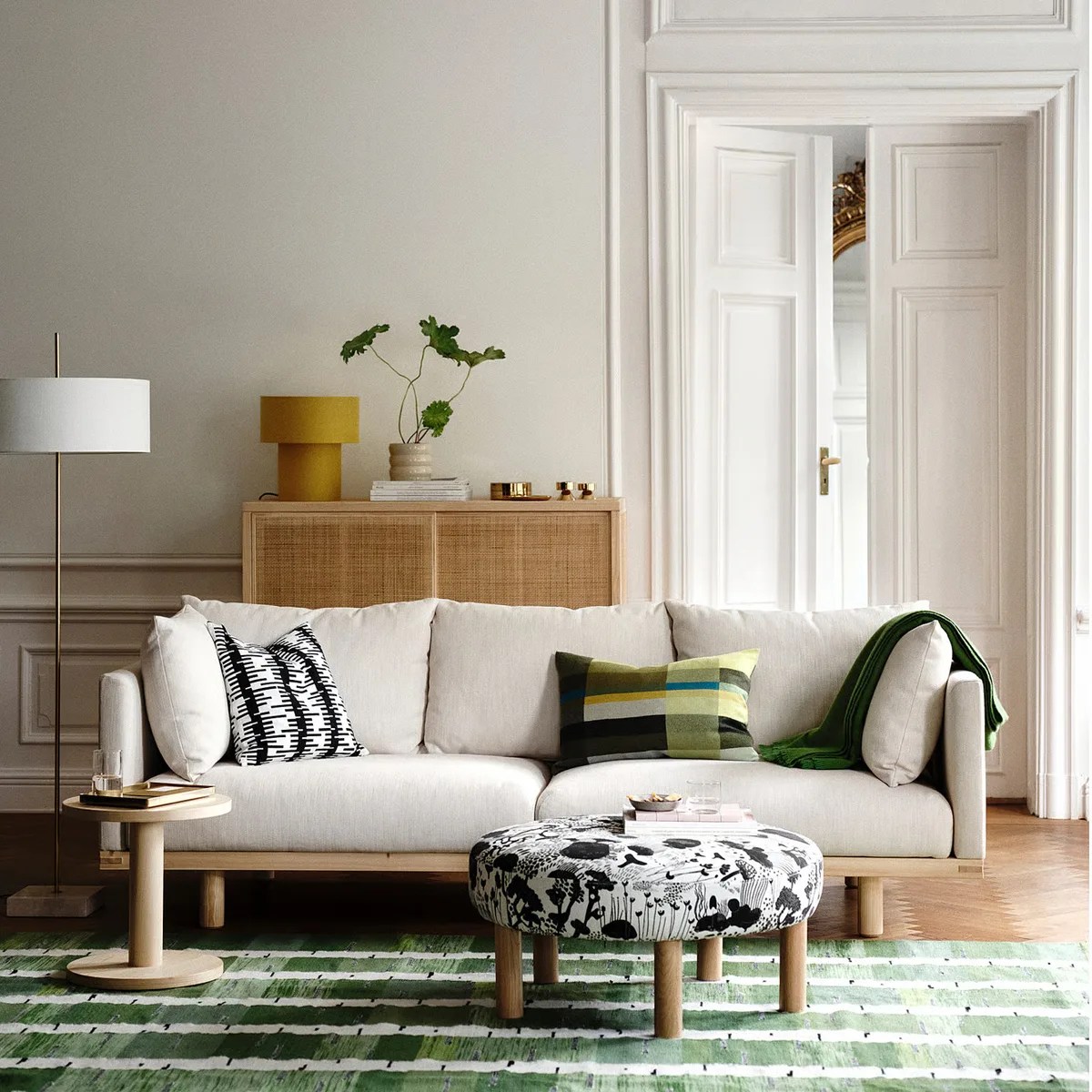 Beige sofa with patterned and green pillows, round black-and-white ottoman, wooden side table, and green striped rug in a bright living room.