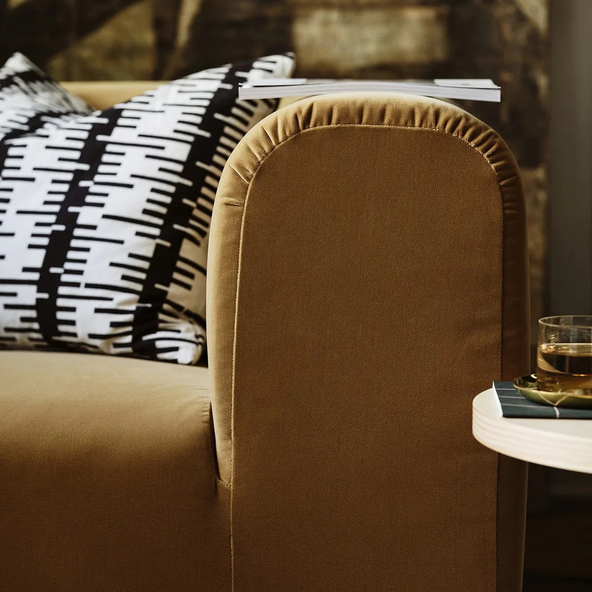 Brown velvet sofa armrest with black and white patterned pillows and a glass on a side table.