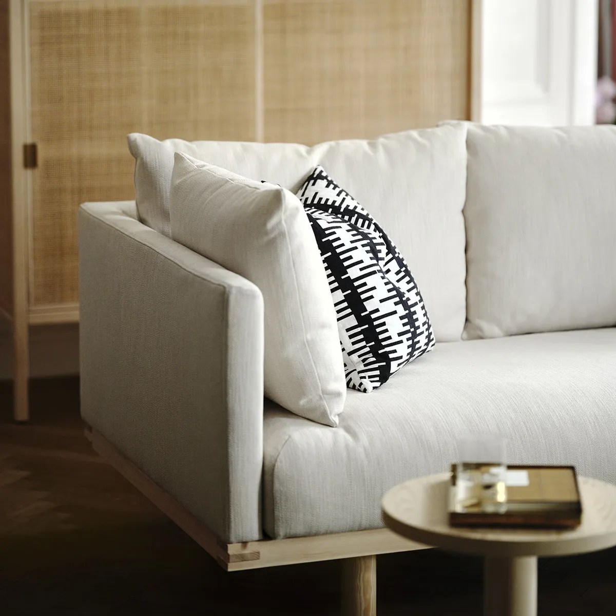 Light beige fabric sofa with wooden base and black-and-white patterned throw pillow.