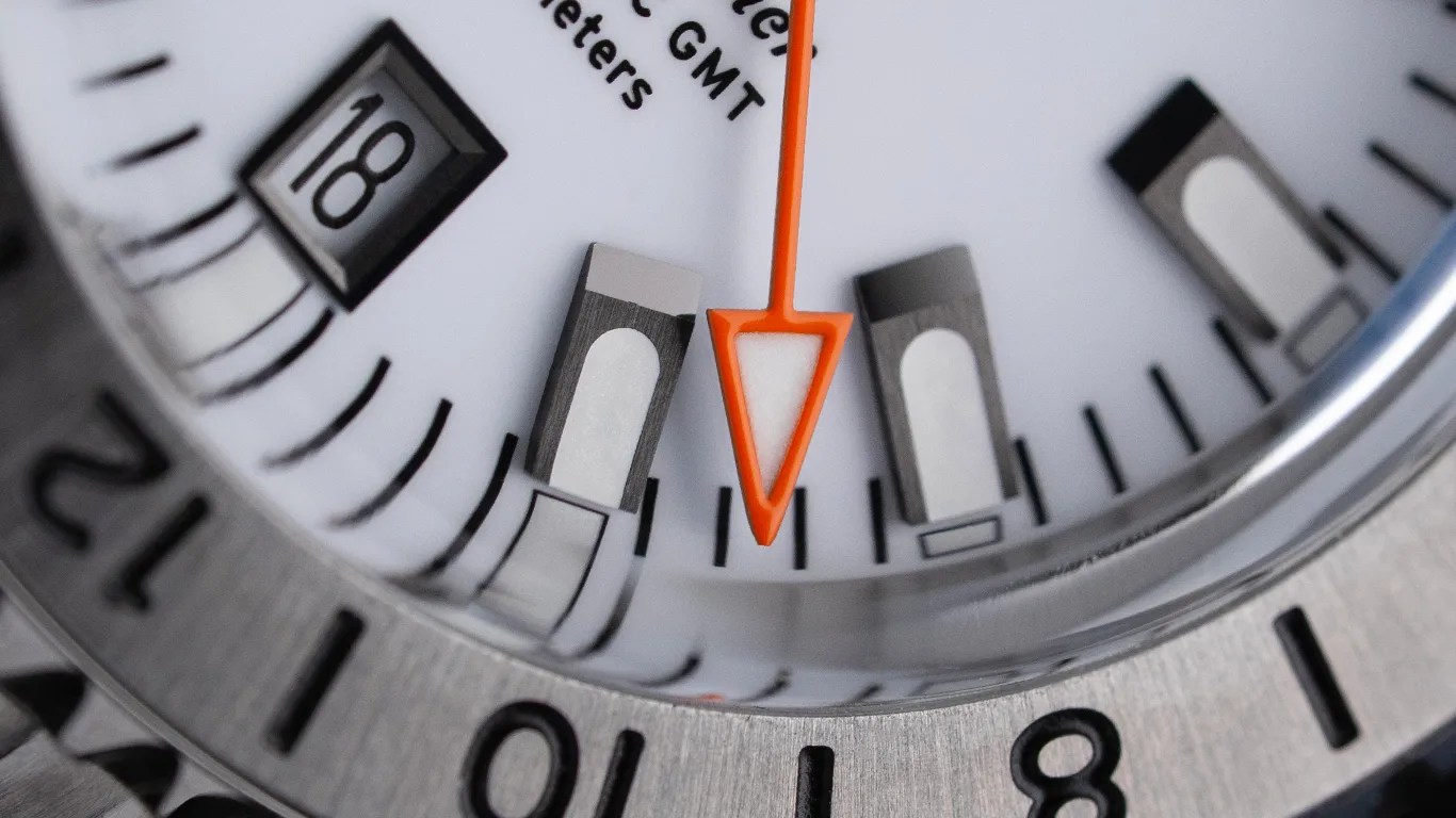 Close-up of a white watch face showing the date "18," black hour markers, and an orange arrow-shaped hand.