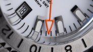 Close-up of a white watch face showing the date "18," black hour markers, and an orange arrow-shaped hand.