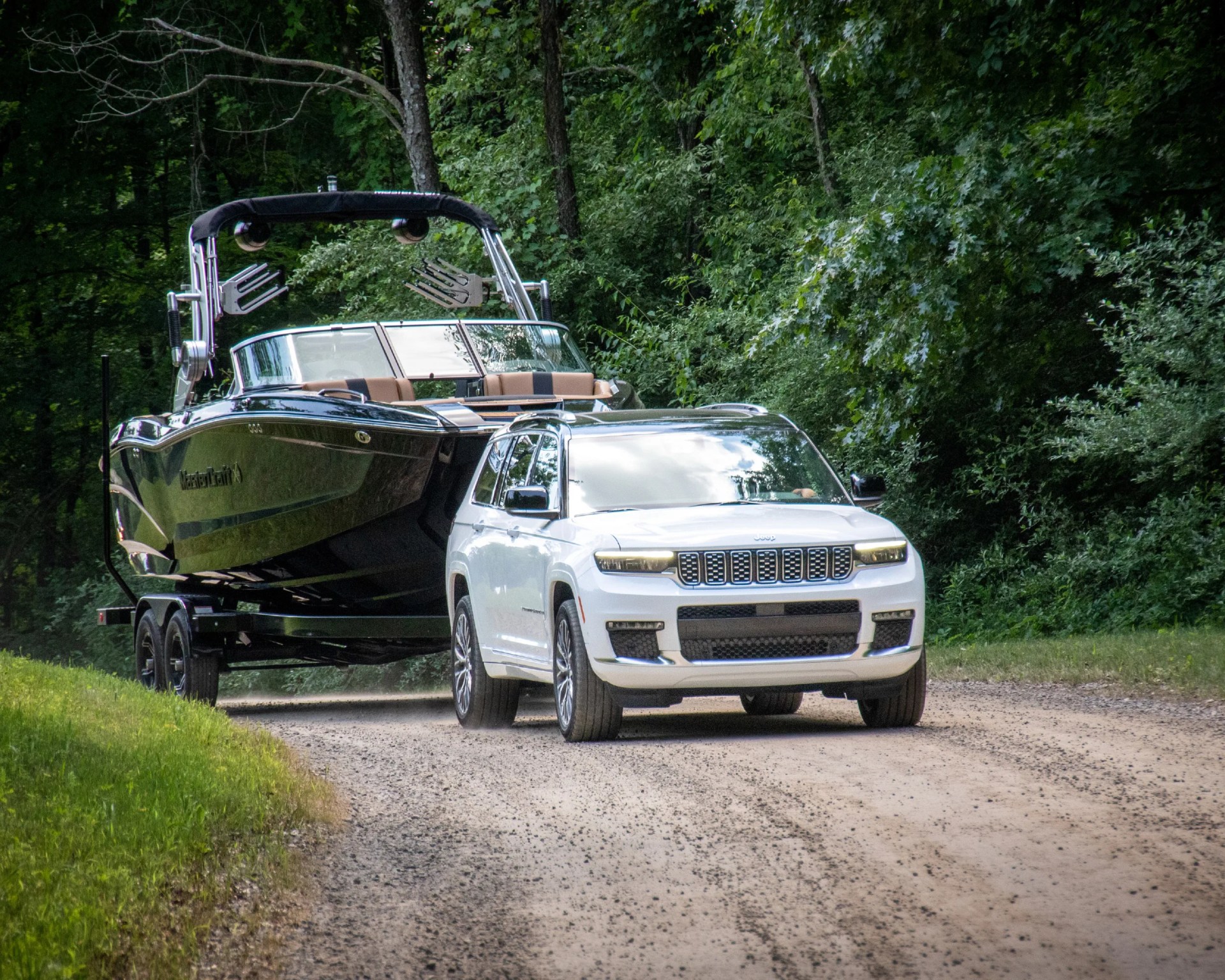 White Jeep SUV towing a black MasterCraft boat on a trailer on a gravel road surrounded by trees.