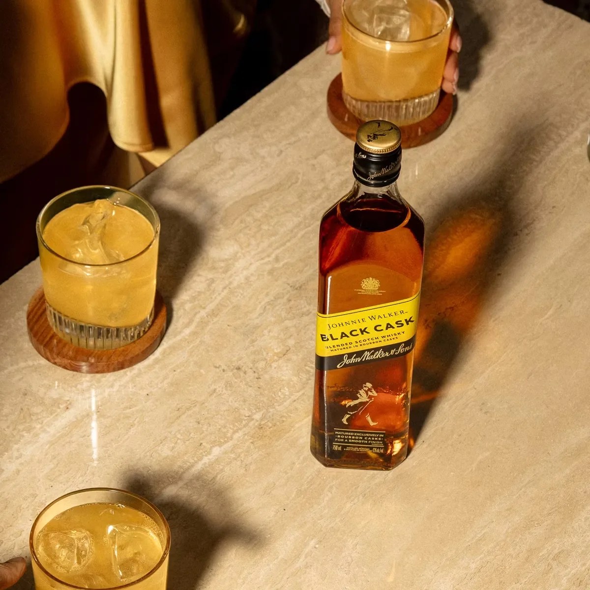 Bottle of Johnnie Walker Black Cask whisky on a marble table with three glasses of whisky on wooden coasters.