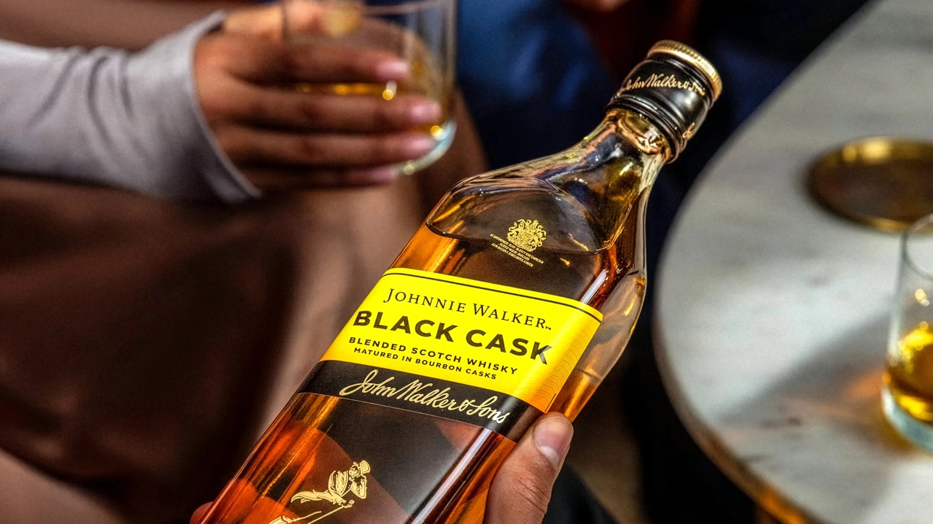Hand holding a bottle of Johnnie Walker Black Cask blended Scotch whisky near a marble table with a glass of whisky.