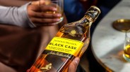 Hand holding a bottle of Johnnie Walker Black Cask blended Scotch whisky near a marble table with a glass of whisky.
