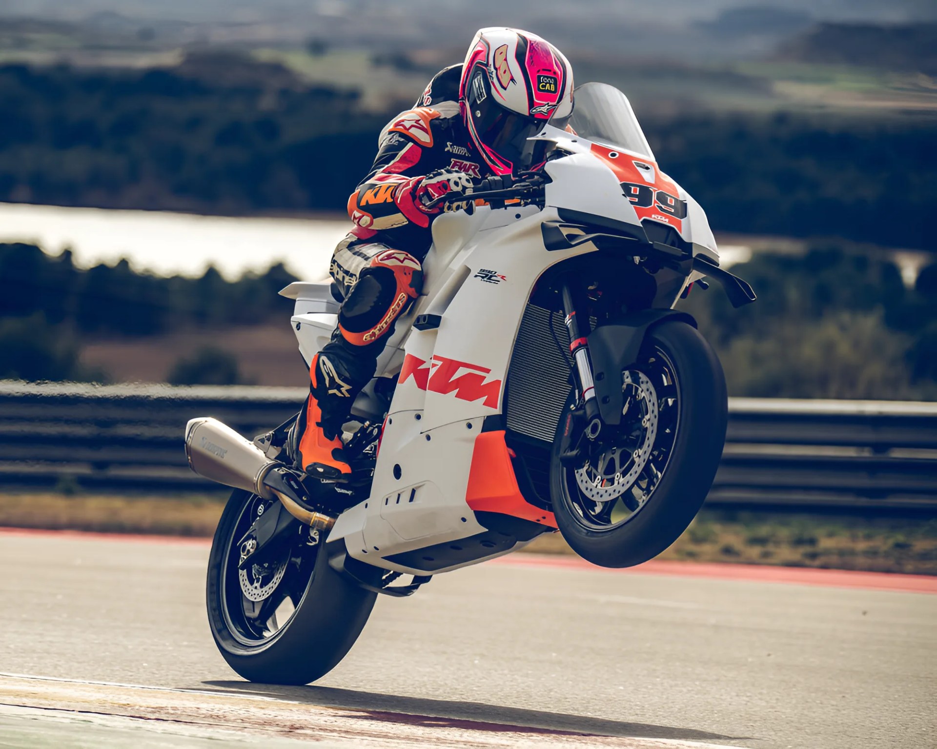 Motorcycle racer in black and orange gear performing a wheelie on a white KTM race bike on a track.