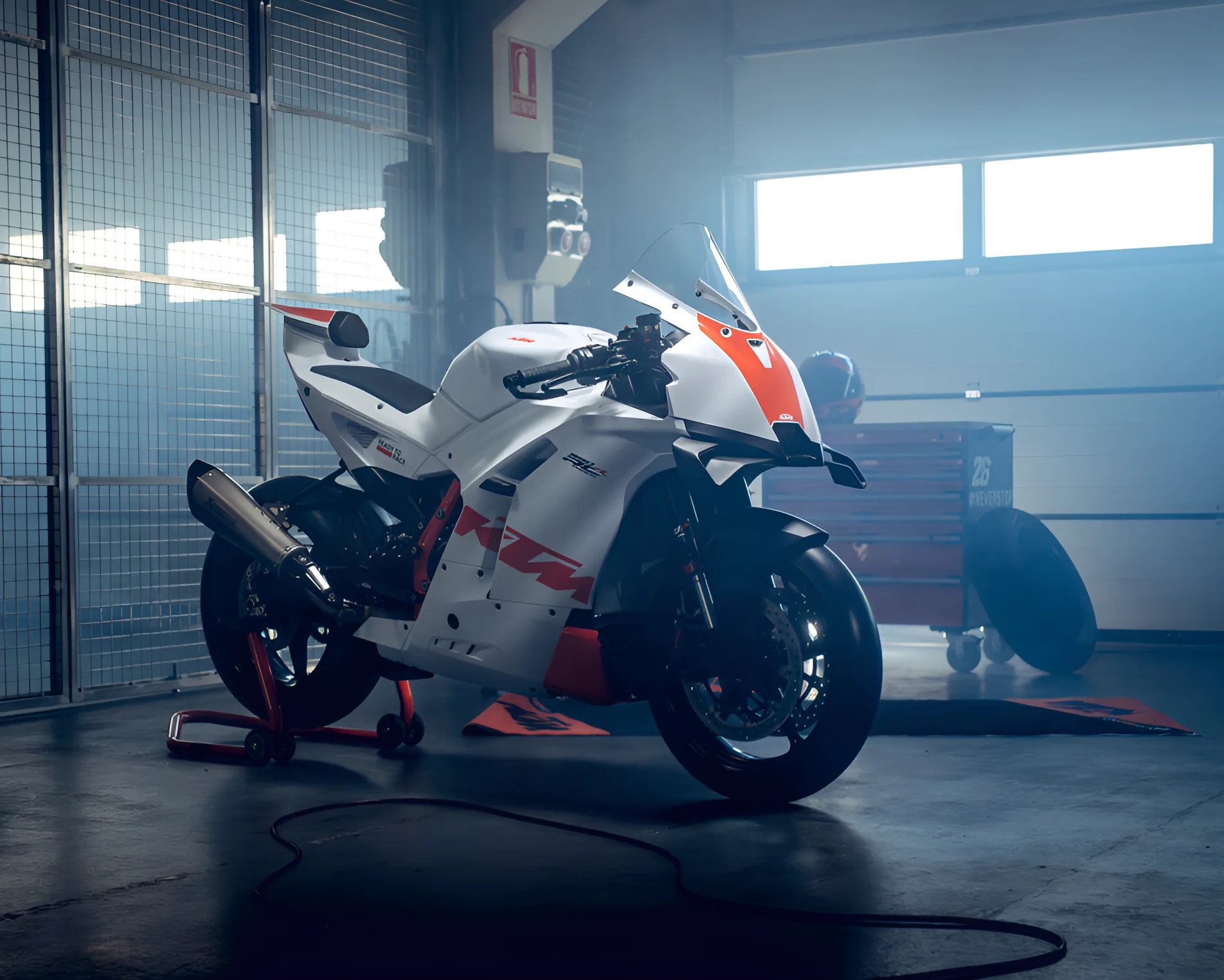 White and orange KTM racing motorcycle on a rear stand inside a garage with a toolbox and helmet in the background.