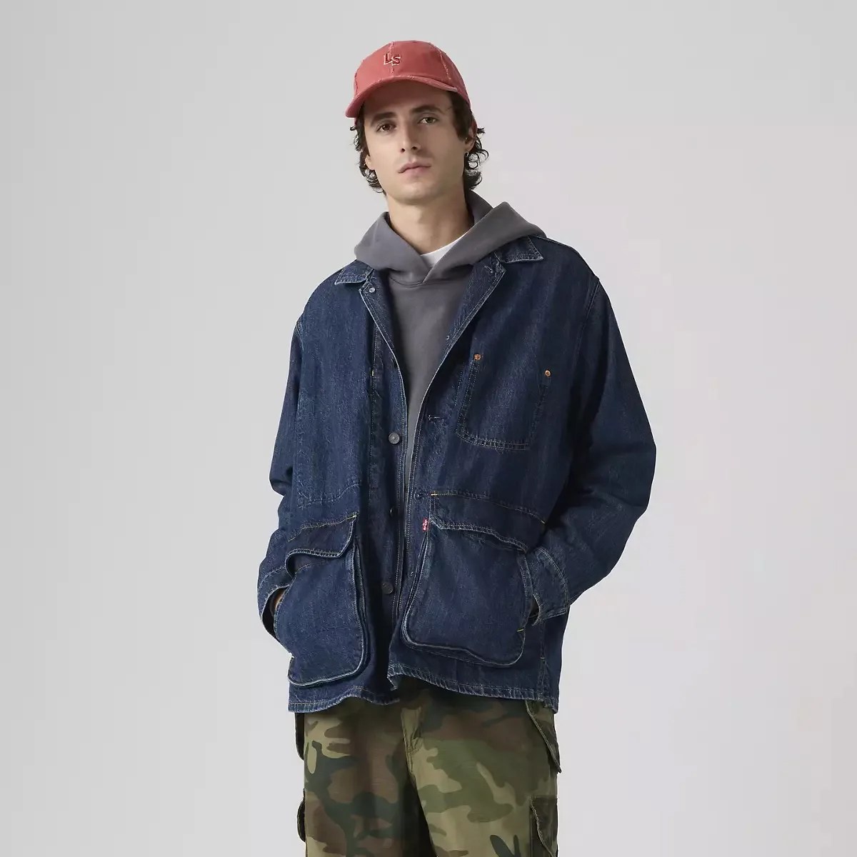 Man wearing a dark blue denim jacket over a gray hoodie, camo pants, and a red cap.