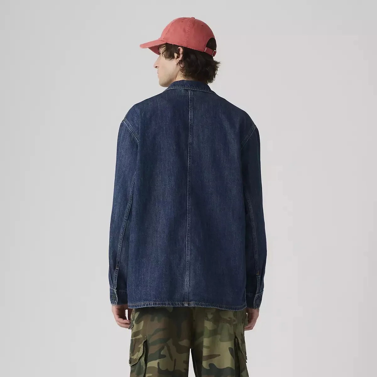 Back view of a person wearing a dark blue denim jacket, camouflage cargo pants, and a red baseball cap.
