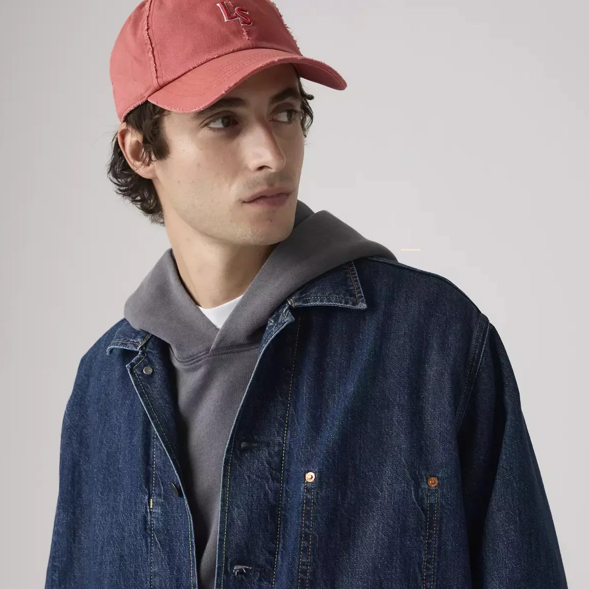 Man wearing a red baseball cap, gray hoodie, and dark blue denim jacket against a plain background.
