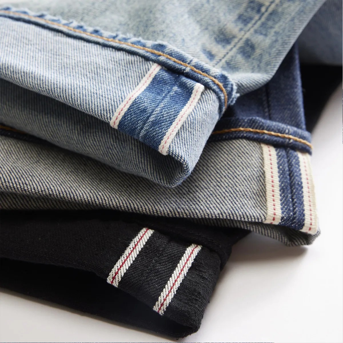 Stack of folded denim jeans in light blue, medium blue, and black with visible selvedge edges.