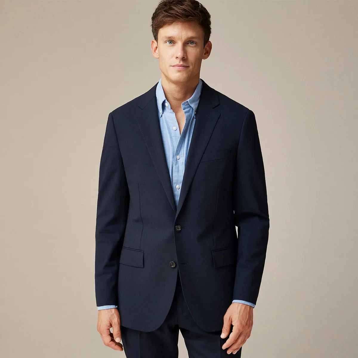 Man wearing a navy blue suit jacket over a light blue button-up shirt against a beige background.