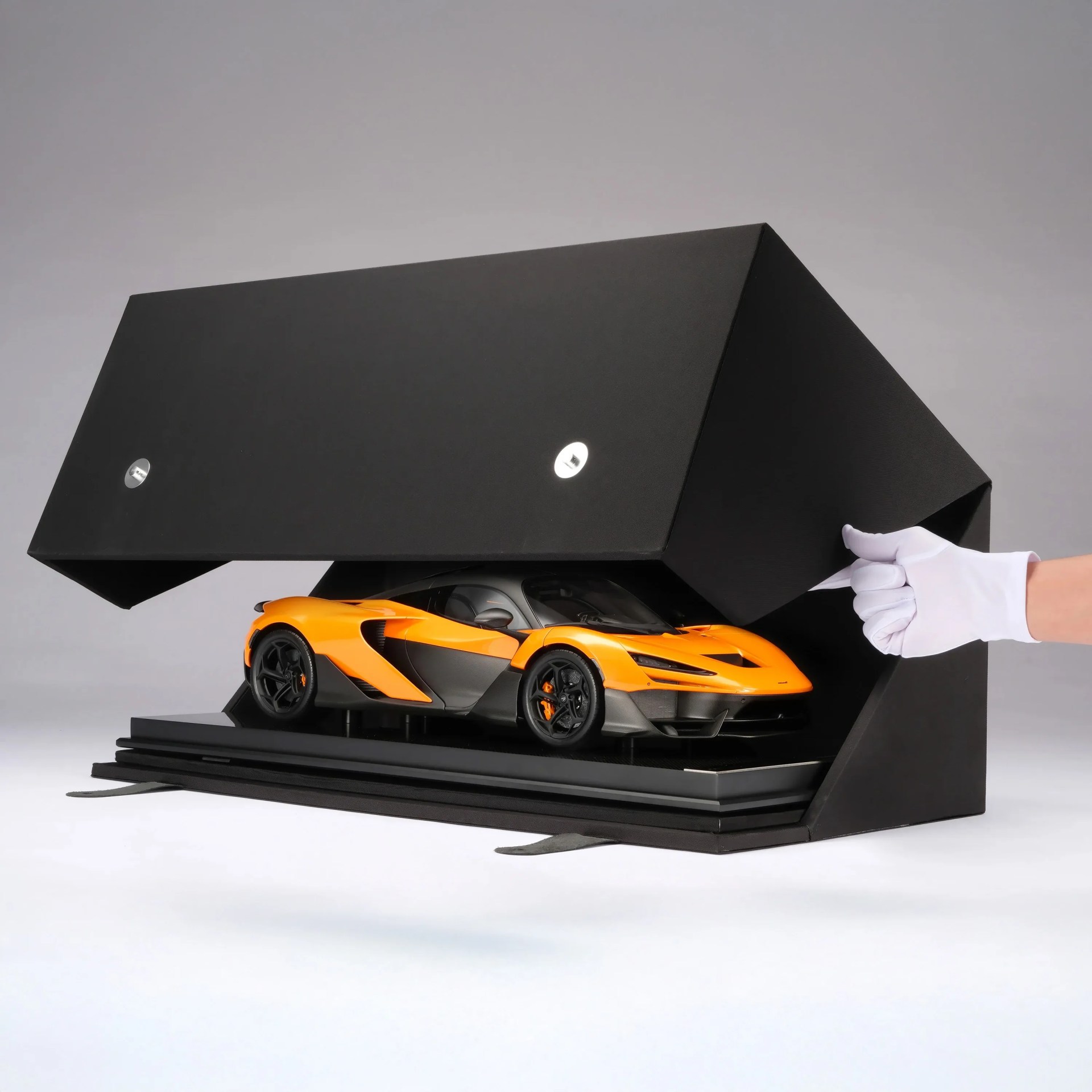 Orange and black sports car model inside a black protective display case being opened by a hand in a white glove.
