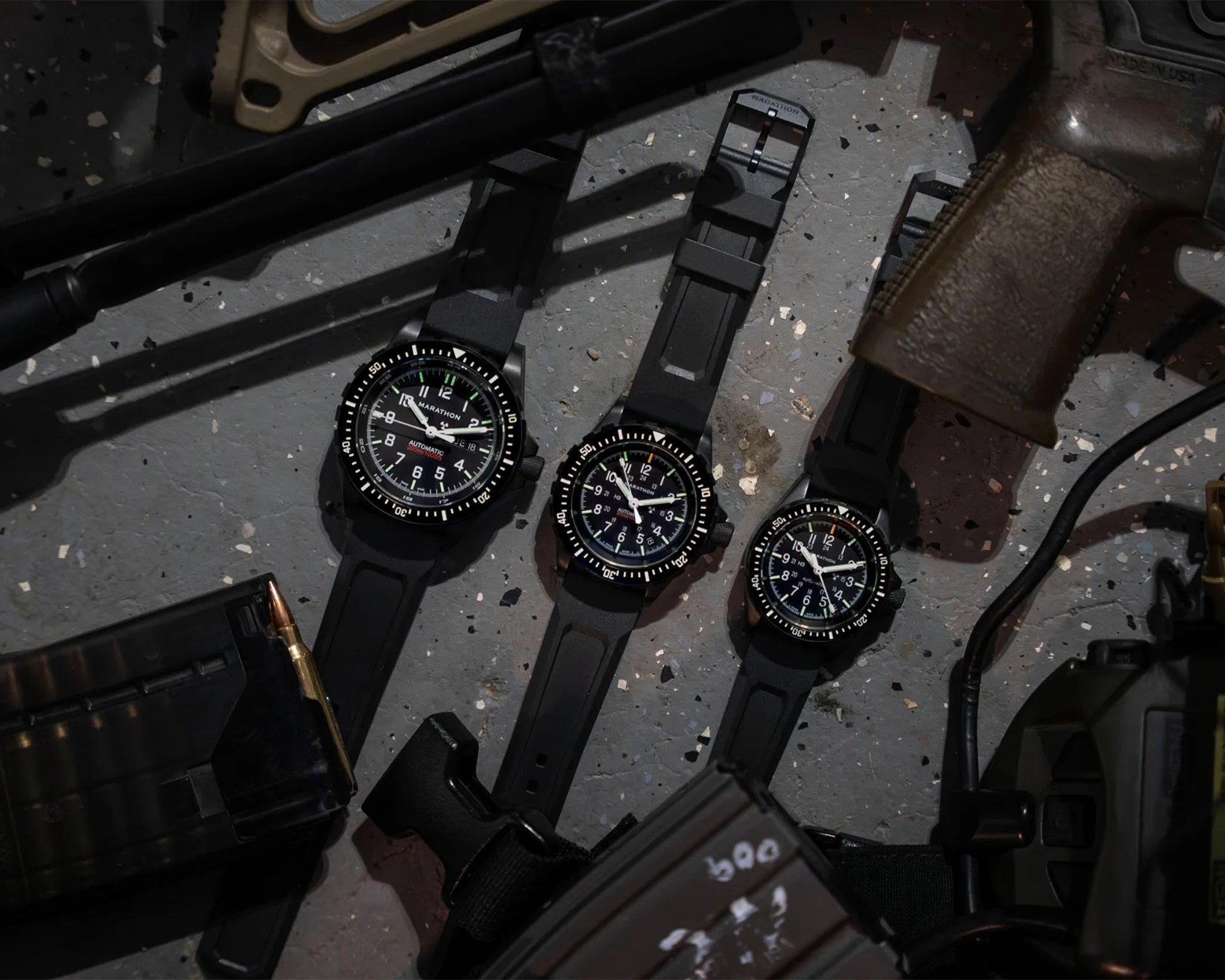 Three black Marathon wristwatches with black rubber straps laid on a speckled gray surface surrounded by firearm parts and ammunition.