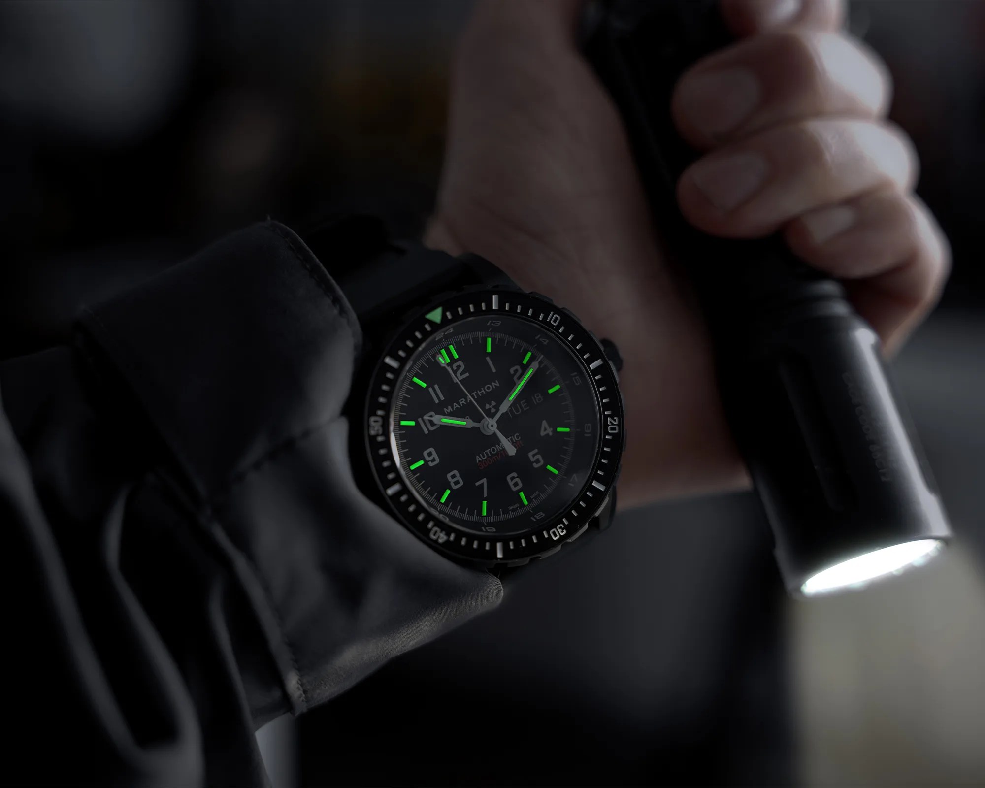 Black Marathon wristwatch with green luminescent hands and markers worn on a wrist holding a flashlight.