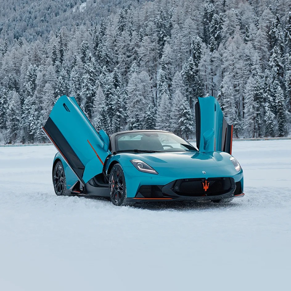 Blue Maserati sports car with butterfly doors open, parked on snow with snow-covered trees in the background.