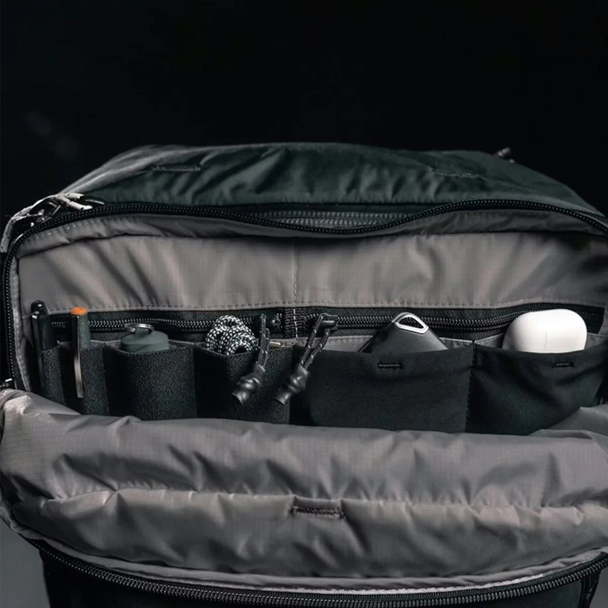 Open black backpack showing multiple small black elastic pockets holding various items including a pen, a small container, a coiled cord, a key fob, and white earbuds case.