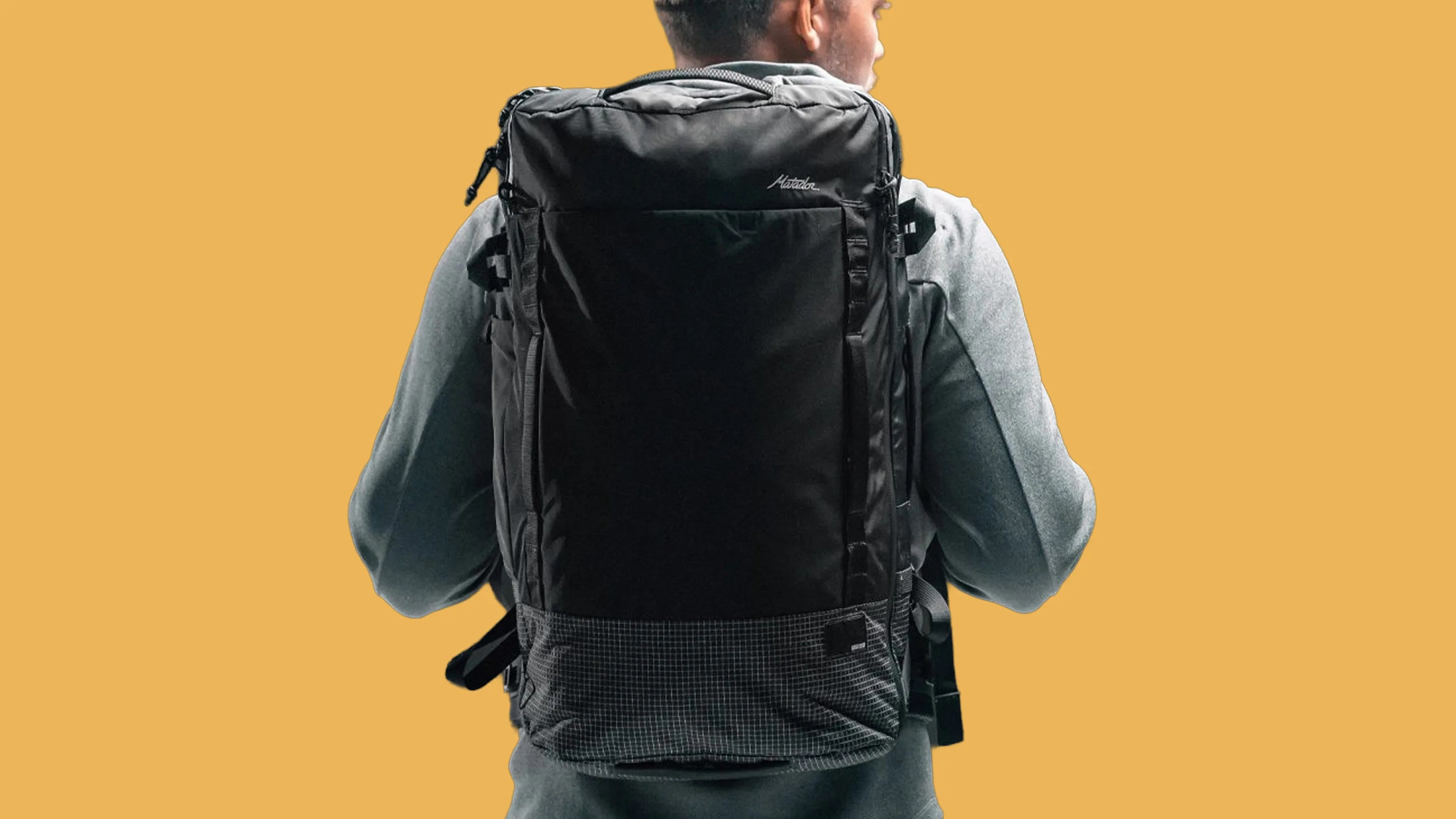 Black rectangular backpack with a grid-patterned bottom worn by a person in a gray jacket.