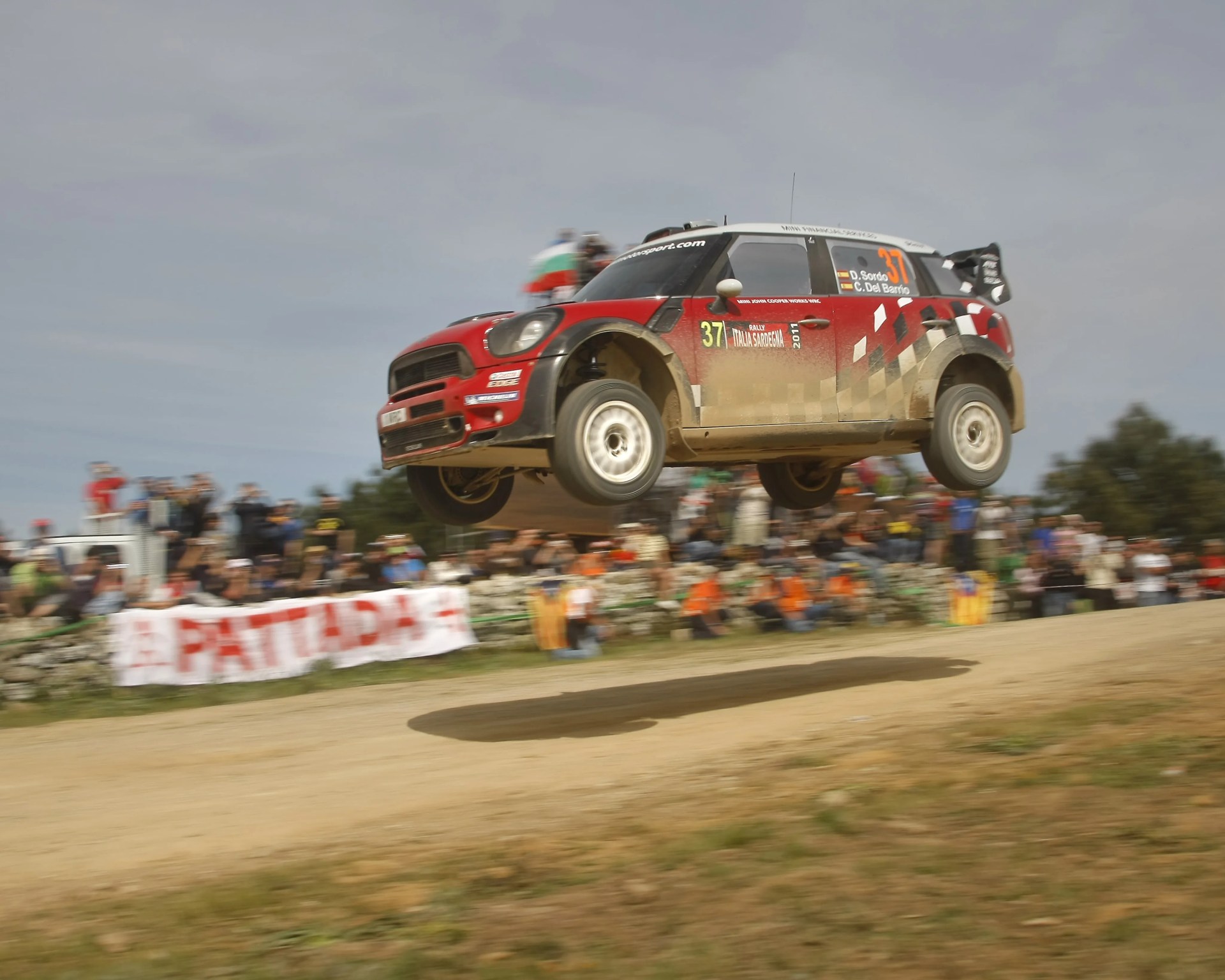 Red and black rally car number 37 airborne on a dirt track with blurred spectators in the background.
