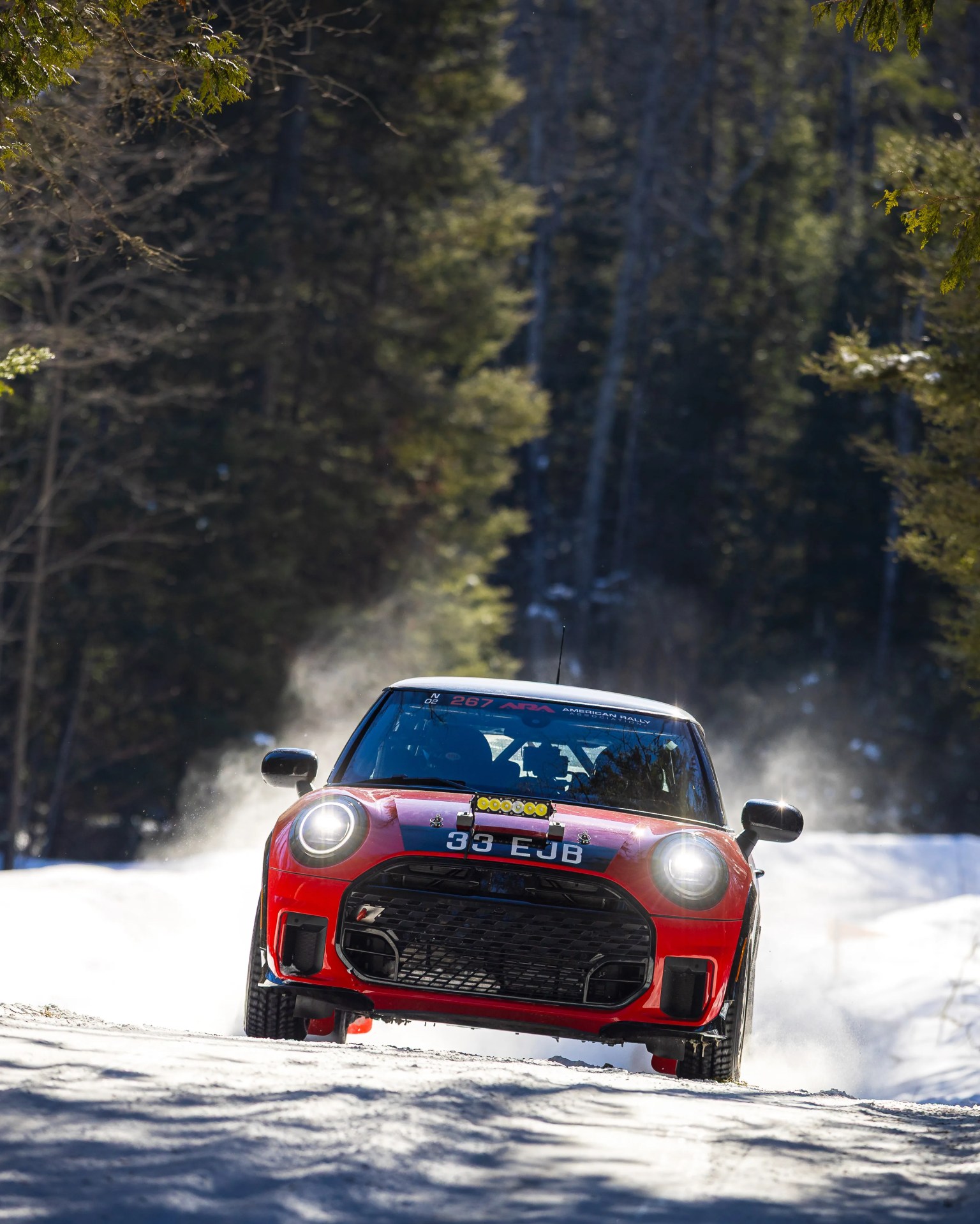 Red rally car with headlights on driving on a snowy forest road.