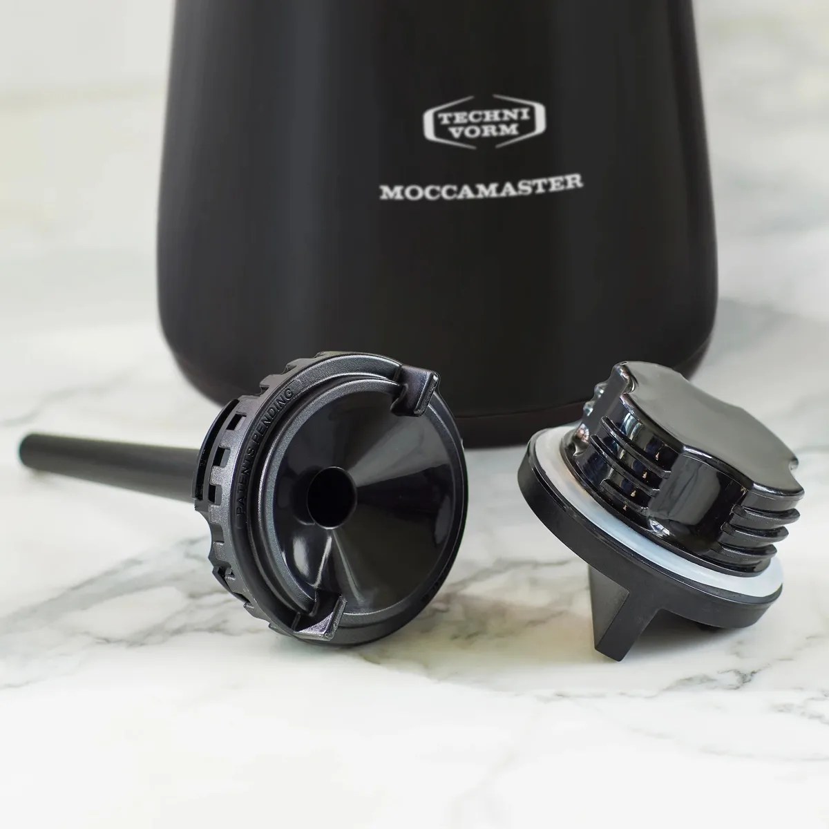 Black plastic coffee maker parts placed on a white marble surface with a black Moccamaster coffee maker in the background.