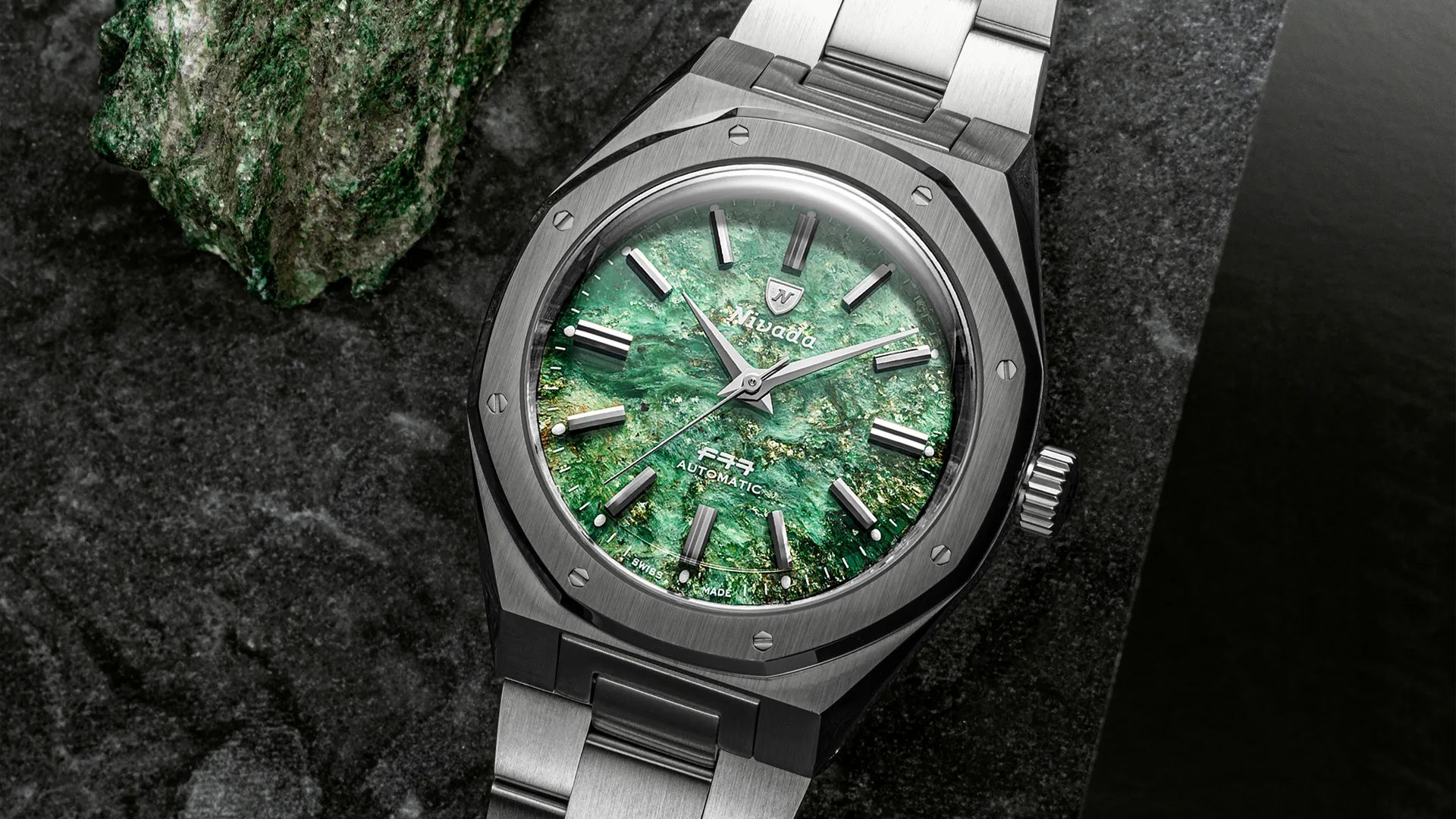 Stainless steel wristwatch with a textured green stone dial and silver hour markers on a dark stone surface.