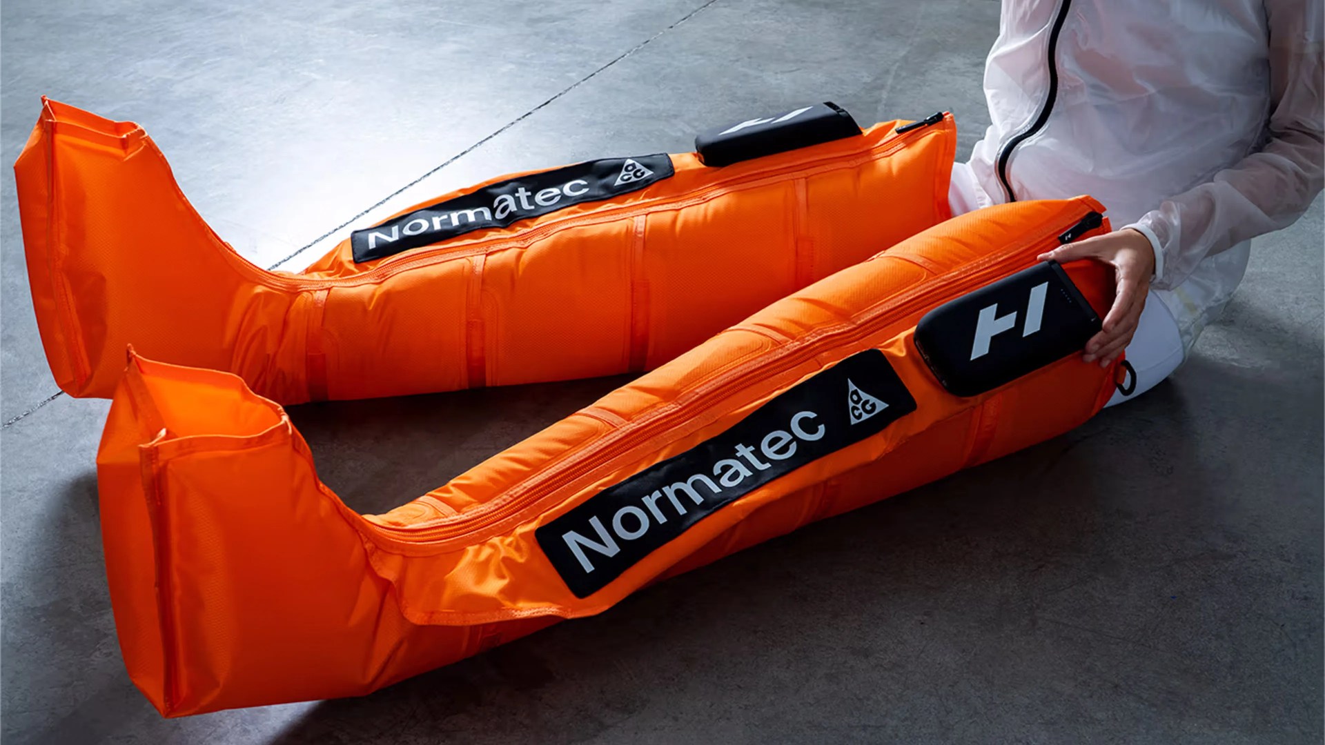 Bright orange Normatec compression leg sleeves worn by a person sitting on a gray floor.