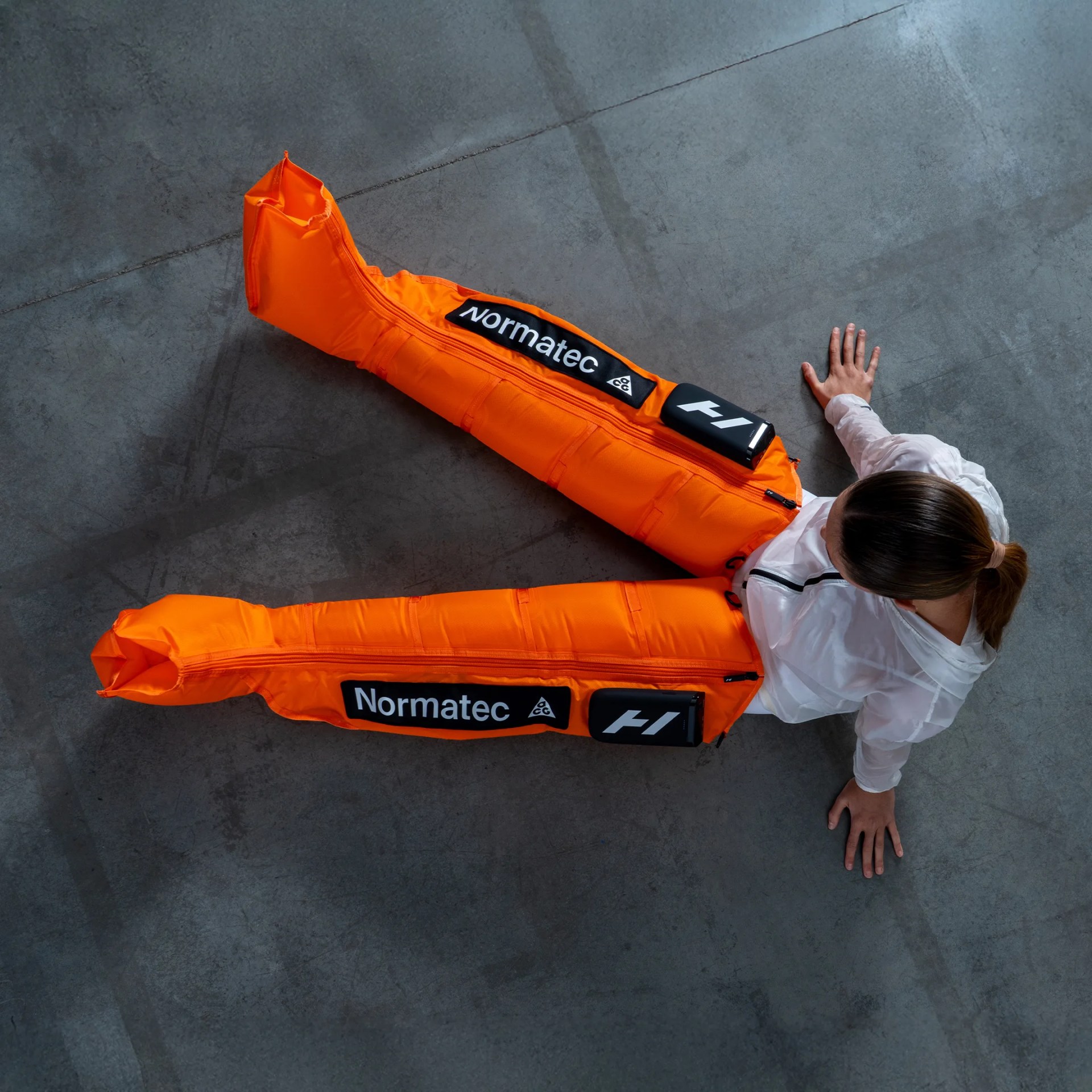 Person wearing bright orange Normatec compression recovery sleeves on both legs, sitting on a gray concrete floor.