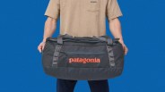 Dark gray Patagonia duffel bag with orange logo held by a person wearing a beige shirt against a blue background.